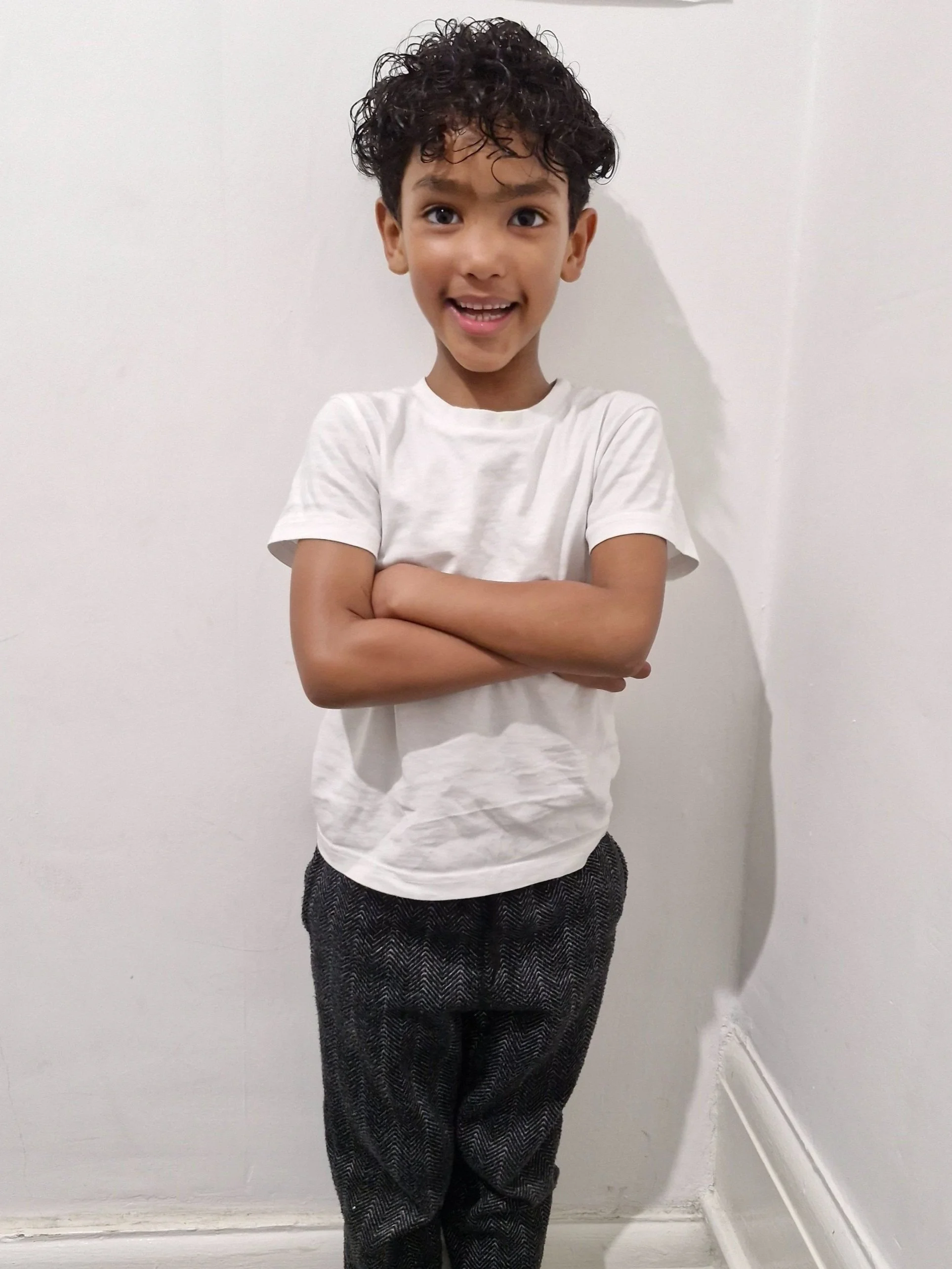 A boy with curly dark hair and brown eyes, smiling with arms crossed, standing against a white wall. Modelling for leading child model and talent agency SocityUK based in London and Manchester.