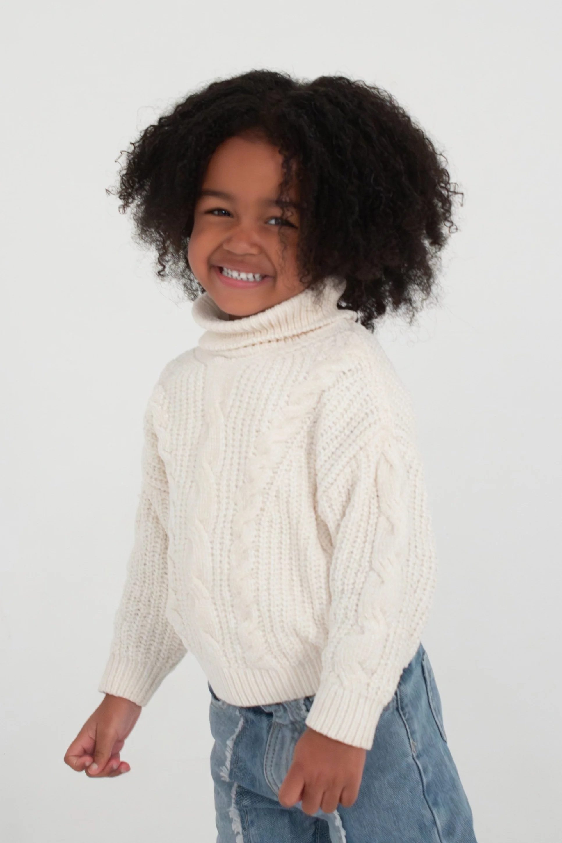 A young girl with curly hair smiling and showing her teeth, wearing a cream-colored turtleneck sweater and baggy jeans, standing against a plain white background. Model for leading Manchester and London based child model agency Socityuk.