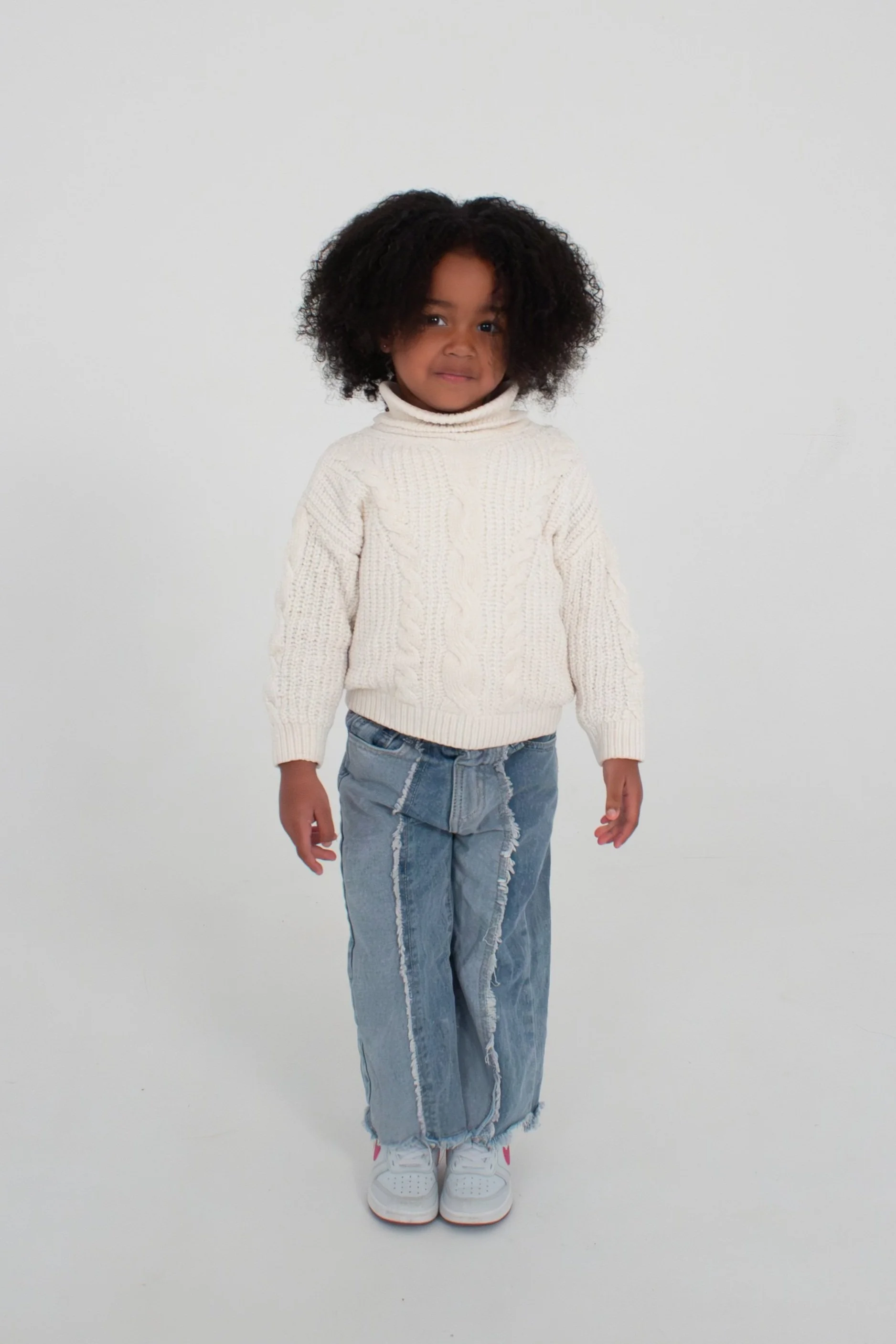 A young girl standing on a white background, wearing a cream-colored cable knit turtleneck sweater, distressed wide-leg jeans, and white sneakers with pink accents. Model for leading Manchester and London based child model agency Socityuk.