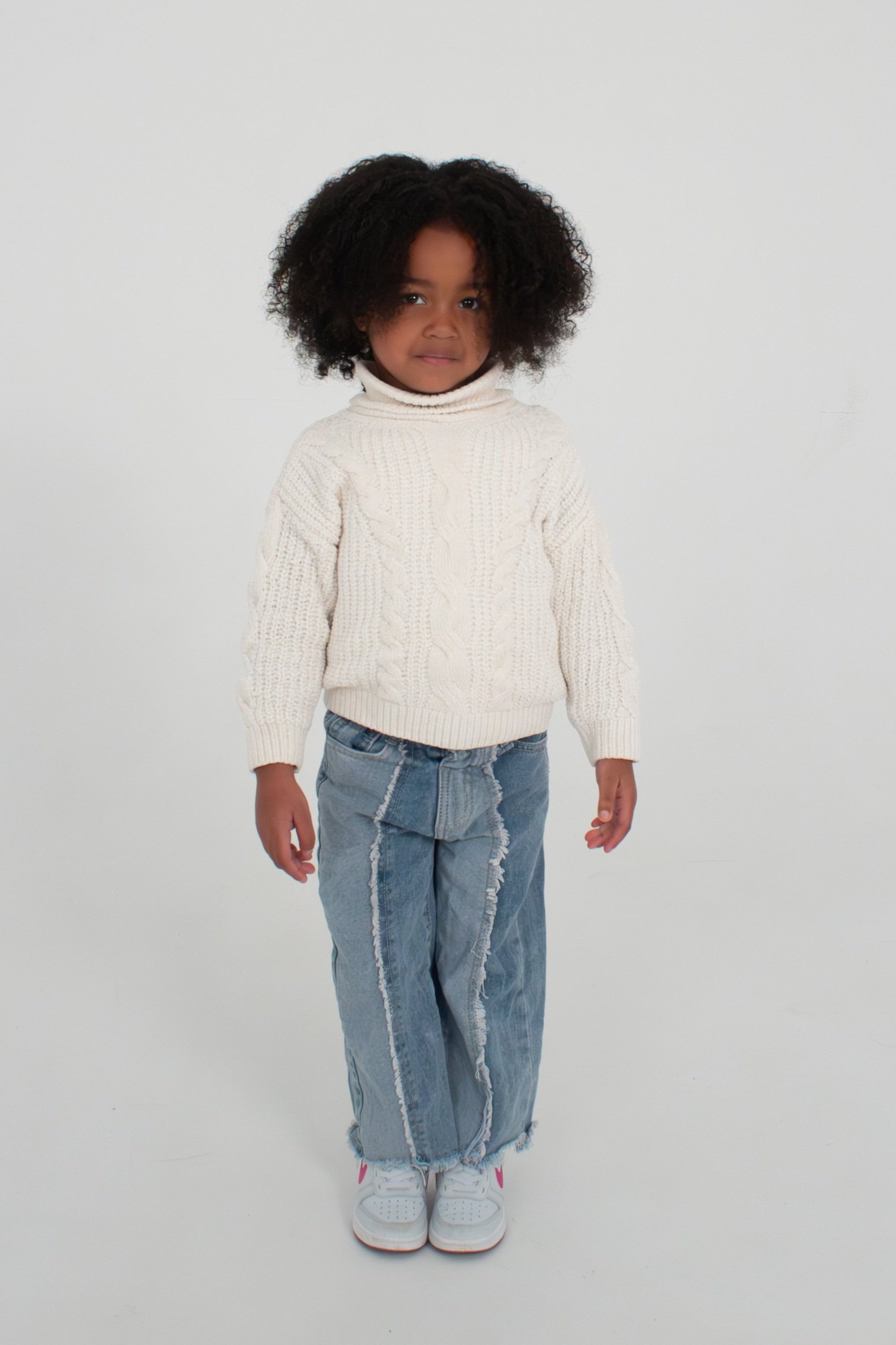 Young girl with dark curly hair wearing a white knit sweater, distressed denim jeans, and white sneakers with pink accents, standing against a plain white background. Model for leading Manchester and London based child model agency Socityuk.