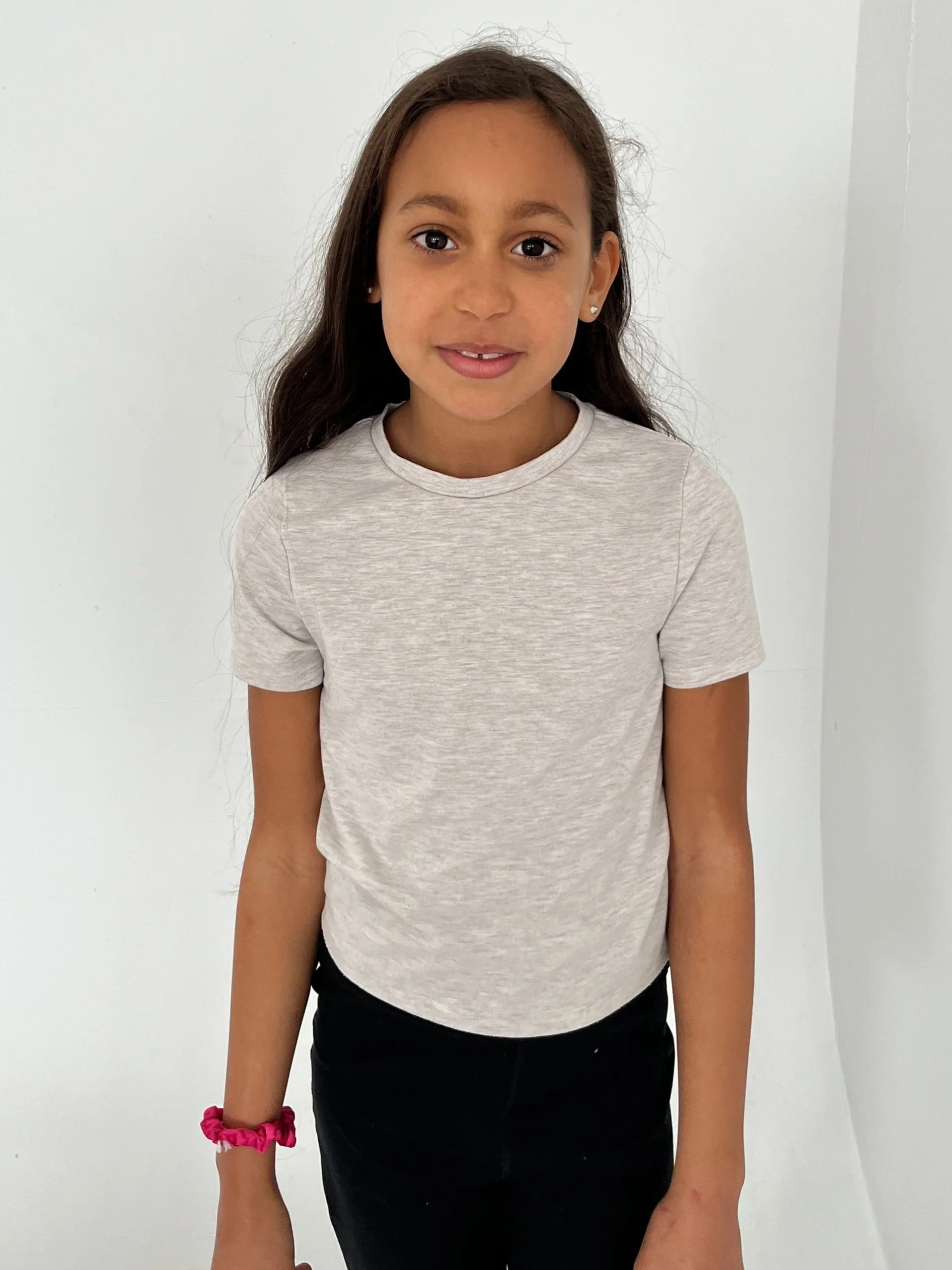 A young girl with long dark hair, wearing a light gray t-shirt, black pants, and a pink scrunchie on her wrist, standing against a plain white background.