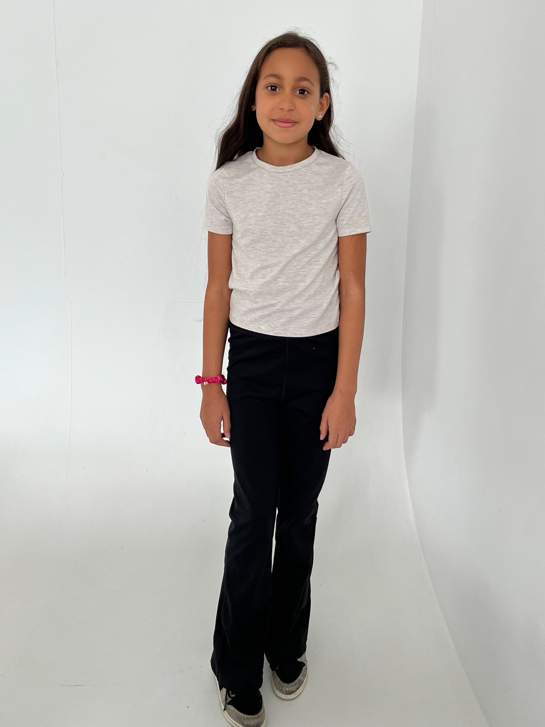 A young girl with long dark hair standing against a plain white background, wearing a light gray t-shirt, black pants, and gray sneakers, looking at the camera with a slight smile.