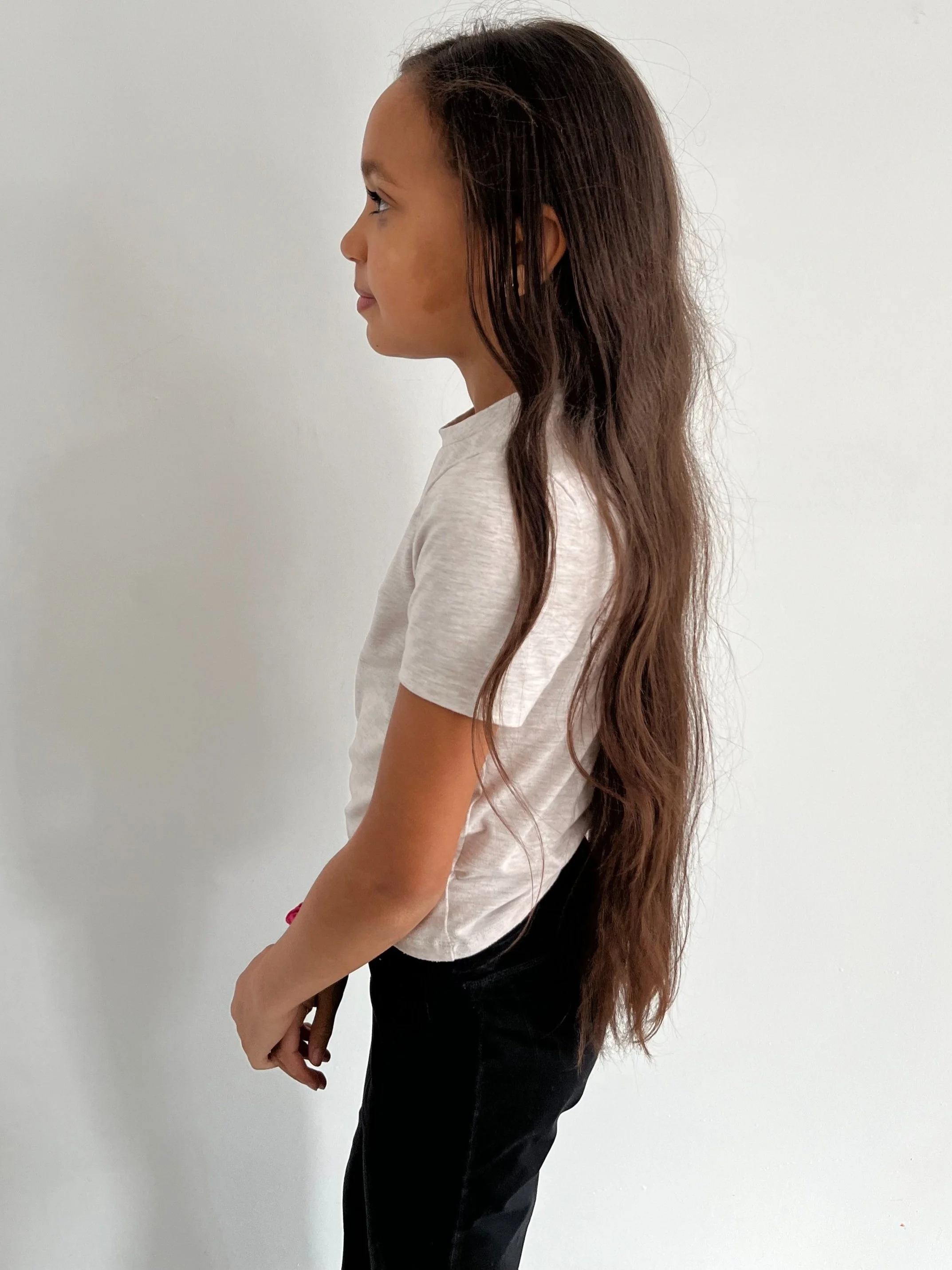 A young girl with long wavy brown hair standing in profile against a plain white wall, wearing a light-colored t-shirt and dark pants. Modelling for leading child model and talent agency SocityUK based in London and Manchester.