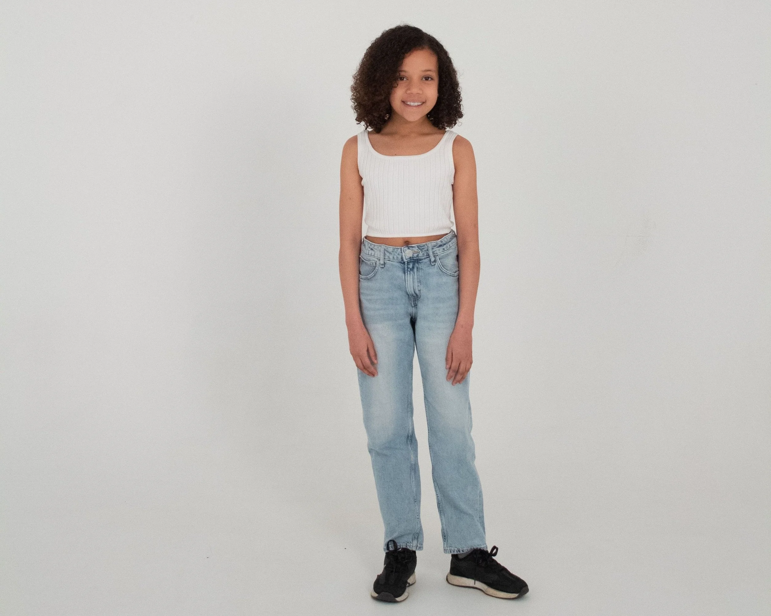 Smiling young girl with curly hair wearing a white sleeveless top, jeans, and sneakers, standing against a plain white background.