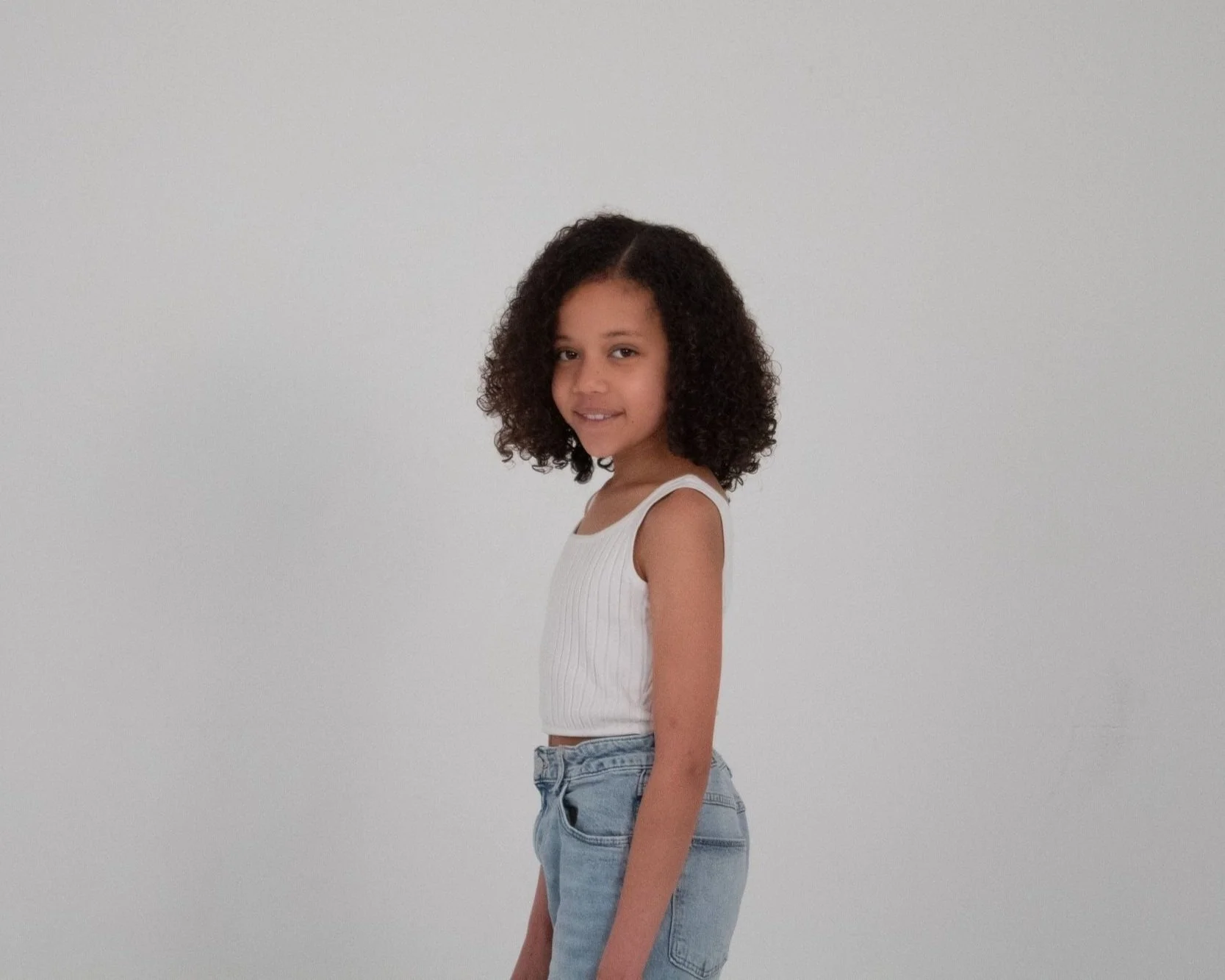 A young girl with curly hair wearing a white sleeveless top and light blue jeans, standing against a plain white background, looking at the camera and smiling.