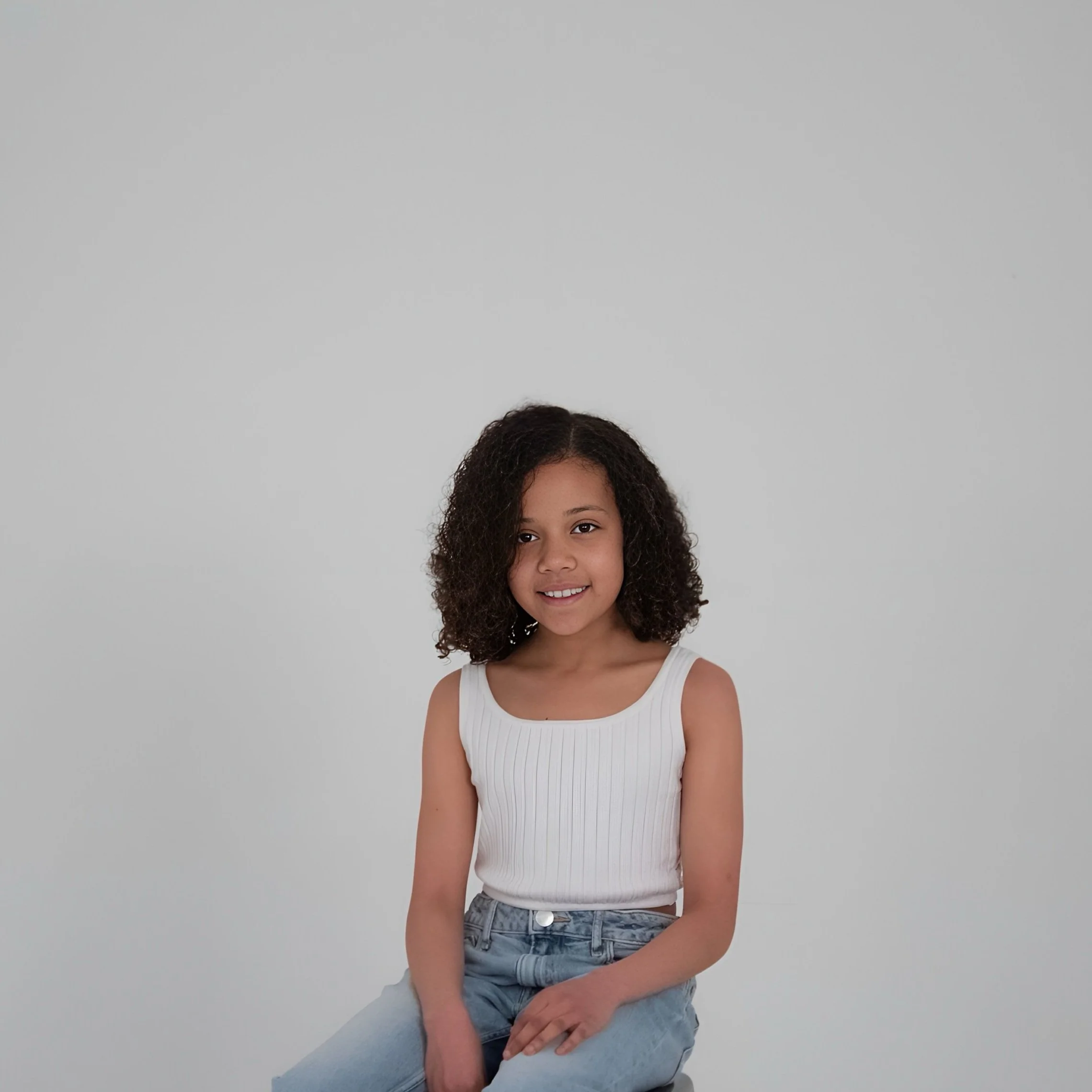 A young girl with curly hair sitting against a plain gray background, wearing a white sleeveless top and light blue jeans, smiling and looking at the camera.