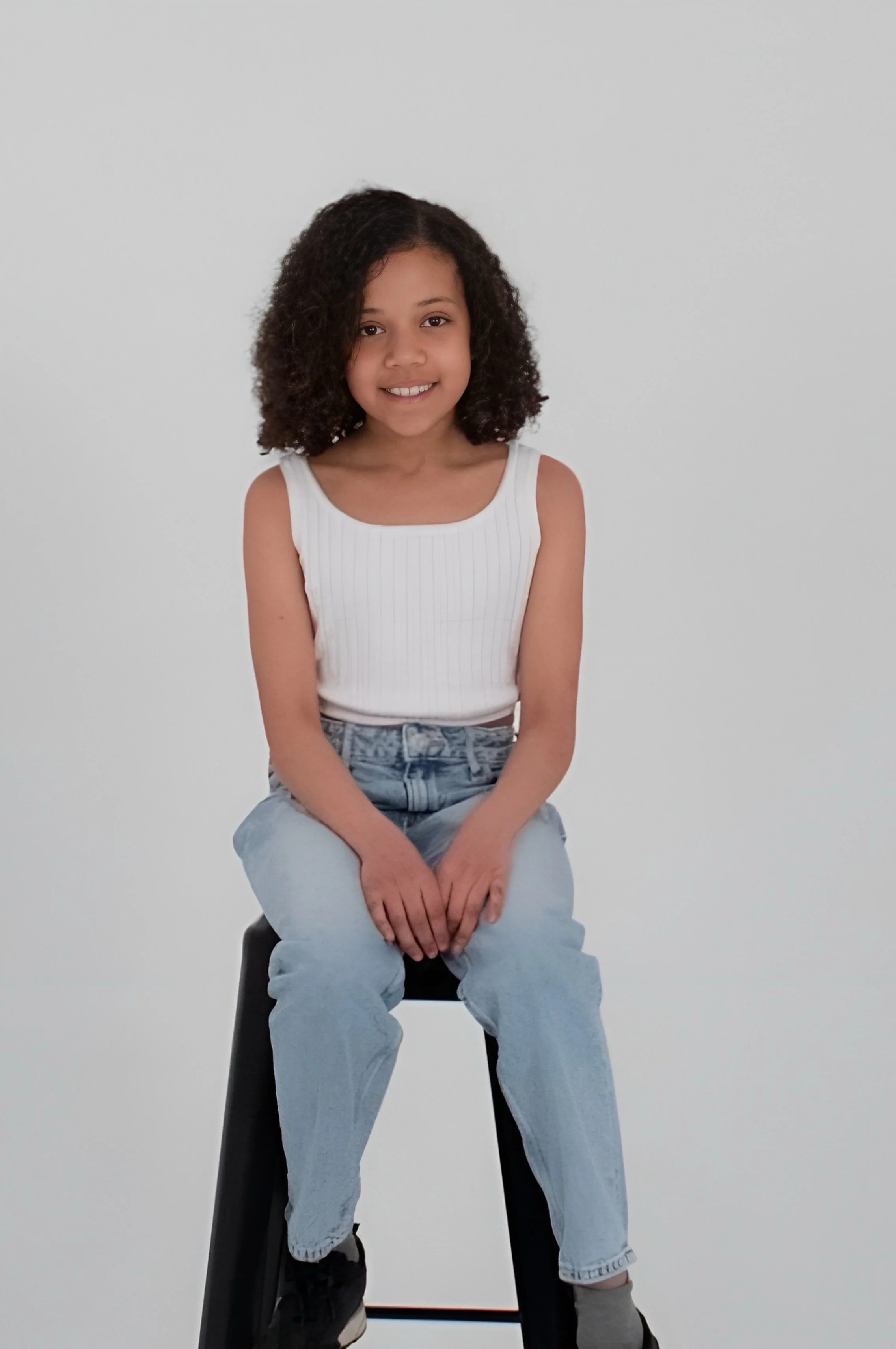 Young girl with curly hair sitting on a black stool against a white background, smiling, wearing a white sleeveless top and light blue jeans.