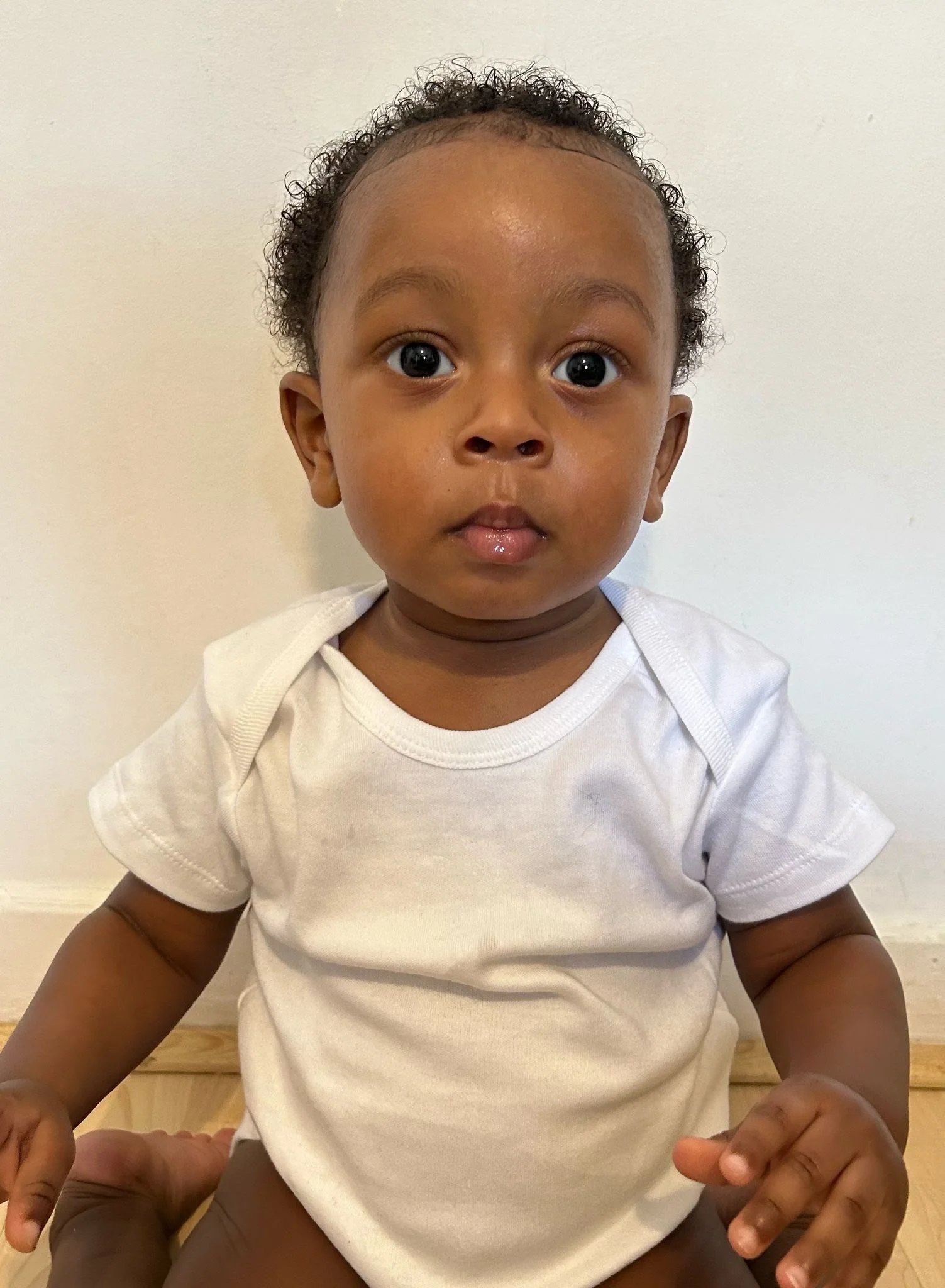 A young child with curly hair and big eyes, wearing a white t-shirt, sitting on a person's lap against a plain white background. Modelling for leading child model and talent agency SocityUK based in London and Manchester.