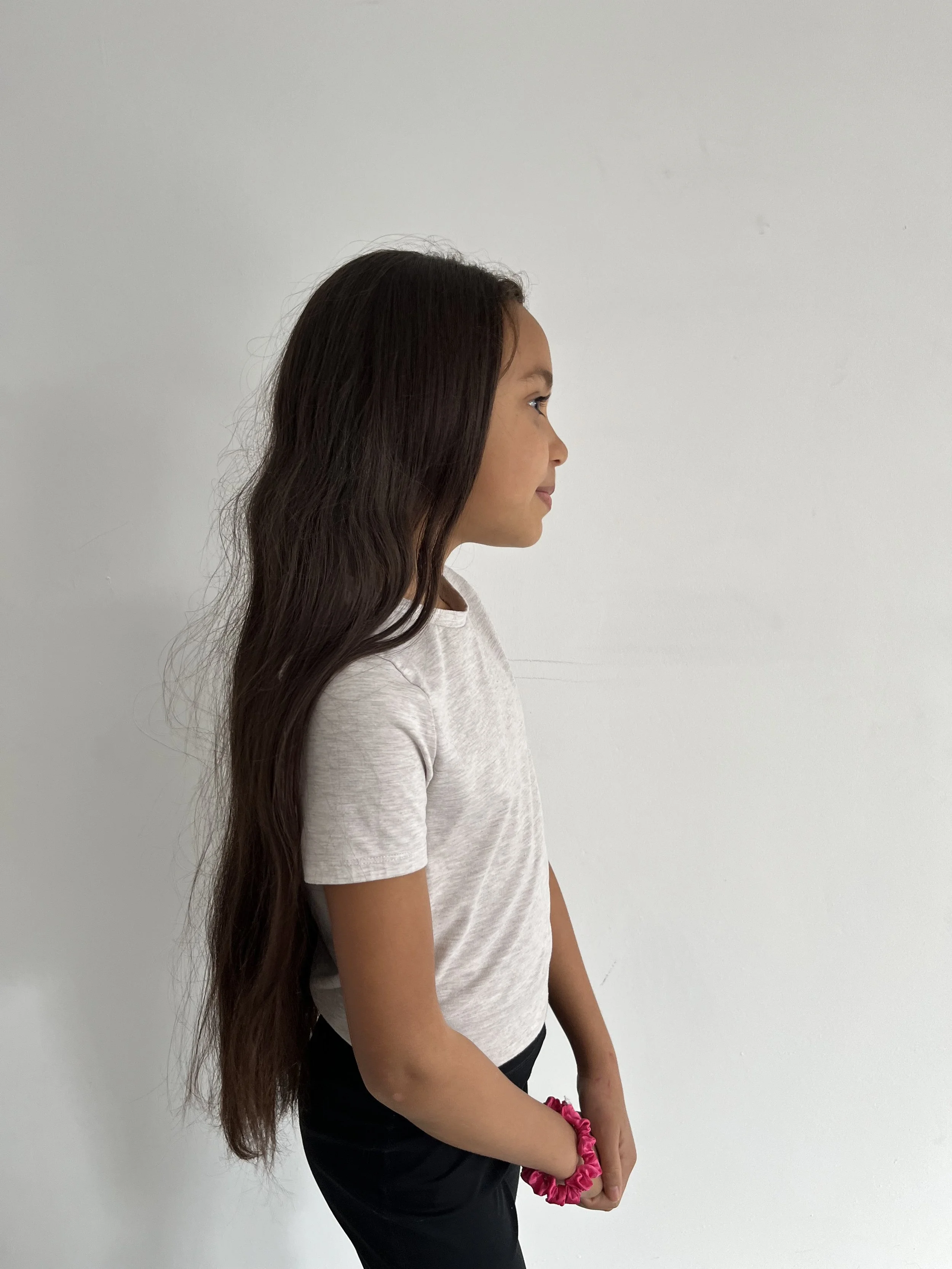 Profile of a young girl with long wavy brown hair wearing a white T-shirt and black pants, standing against a plain white wall.