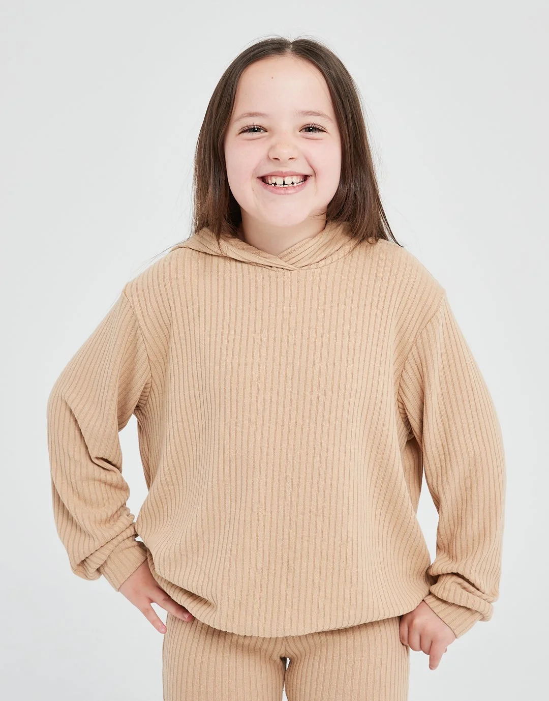 A young girl with brown hair smiling, wearing a matching beige ribbed hoodie and pants, standing against a plain white background.