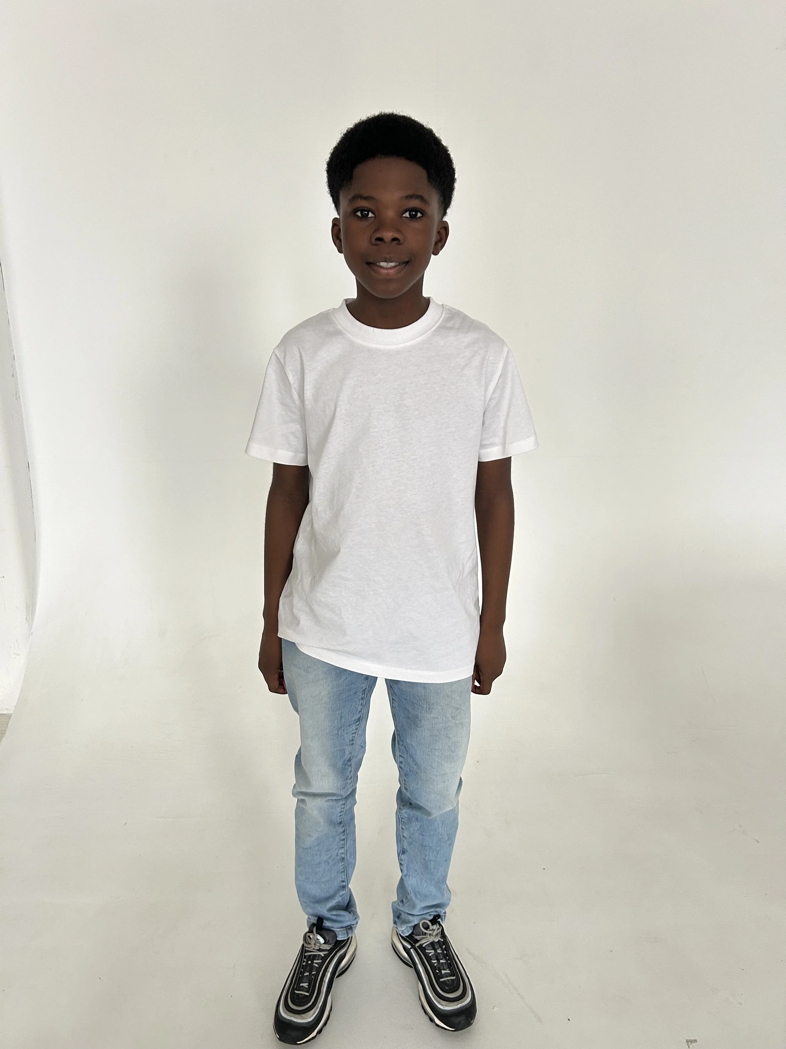 A young boy with dark hair in a fade haircut, wearing a plain white T-shirt, light blue jeans, and stylish sneakers, standing against a white background and smiling at the camera.