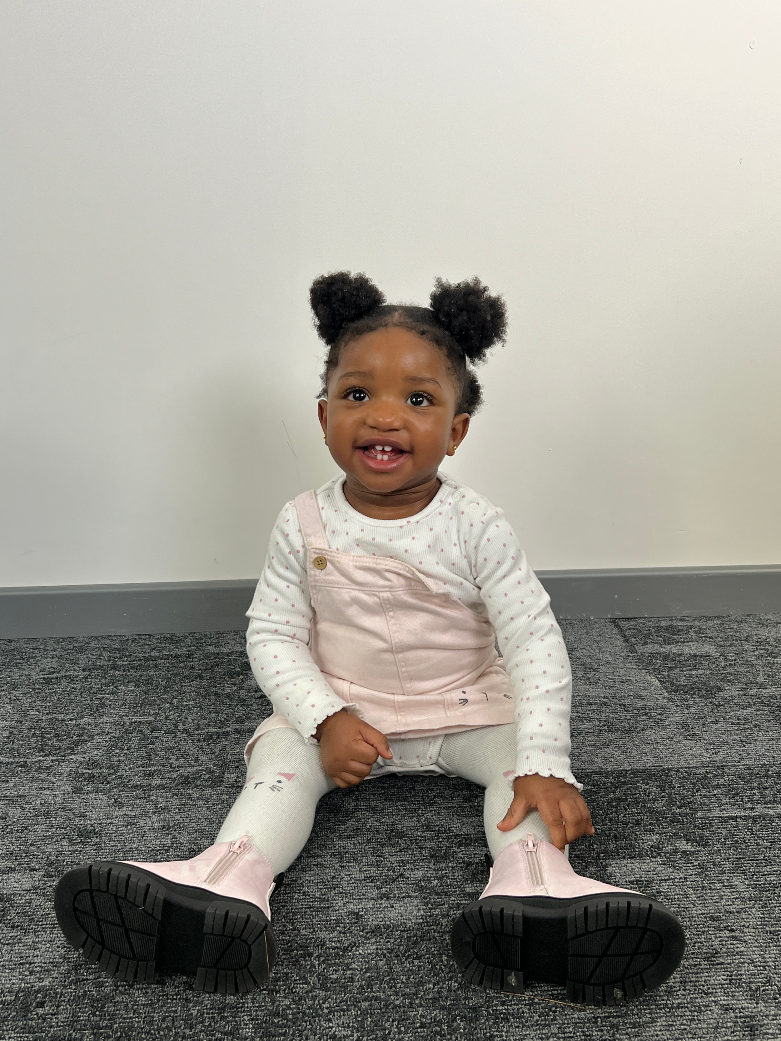 A smiling young girl with dark curly hair styled in two buns, wearing a white polka dot shirt, light pink overalls, white leggings with a small animal face design, and light pink boots, sitting on a gray carpeted floor against a plain white wall.