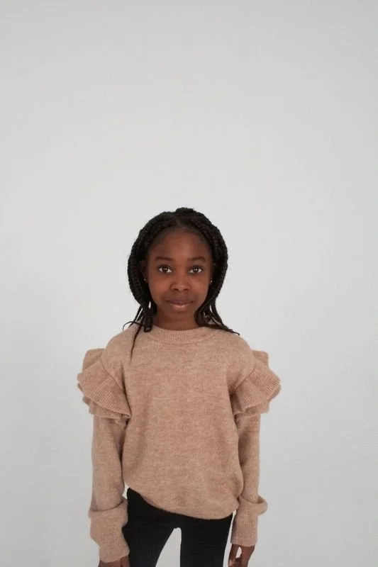 A young girl with braids wearing a beige sweater with ruffled shoulders and black pants, standing against a plain white background.