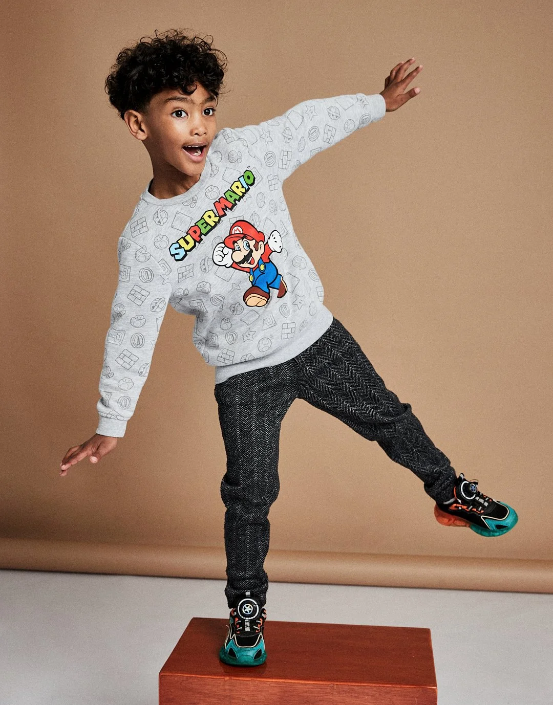 Young boy with curly hair striking a playful pose on a brown platform, wearing a Super Mario sweatshirt, dark patterned pants, and colorful sneakers against a beige background. Modelling for leading child model and talent agency SocityUK based in Lon