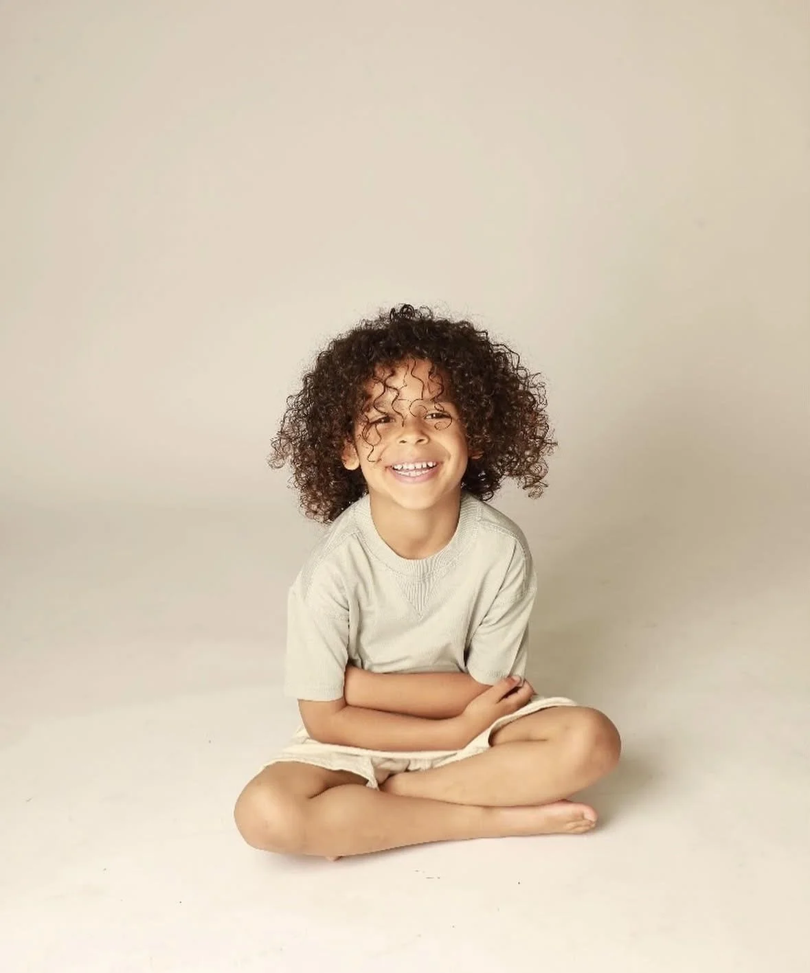 A young boy with curly hair smiling and sitting cross-legged against a plain light background.