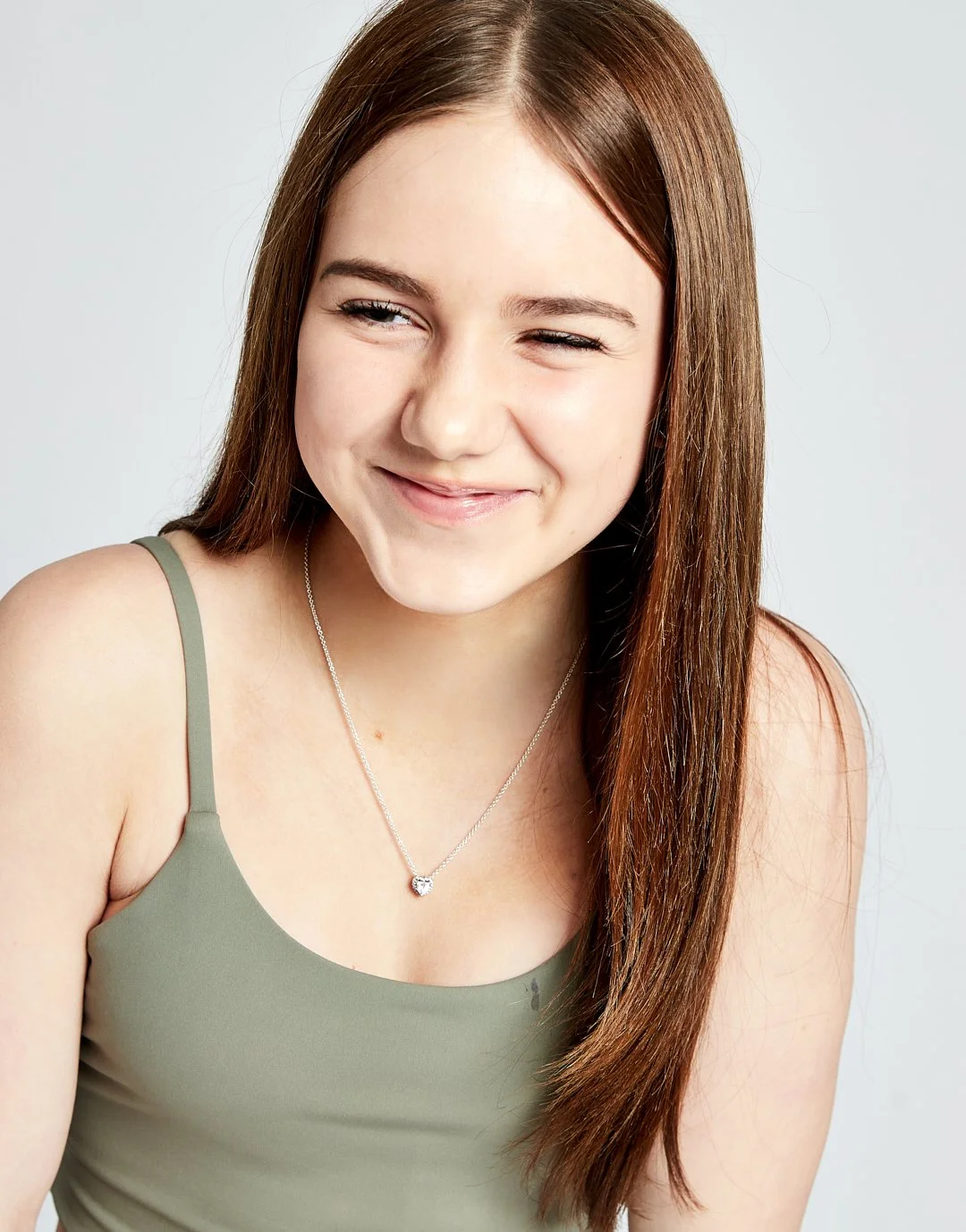 A young woman with long brown hair wearing a green tank top and a silver necklace with a heart-shaped pendant, smiling with her tongue slightly out.