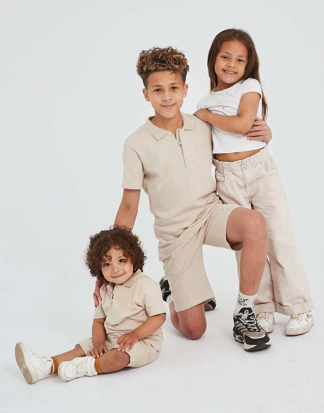 Four children, two boys and two girls, posing together against a plain white background. They are dressed casually in beige and white clothing, with some sitting and others standing.