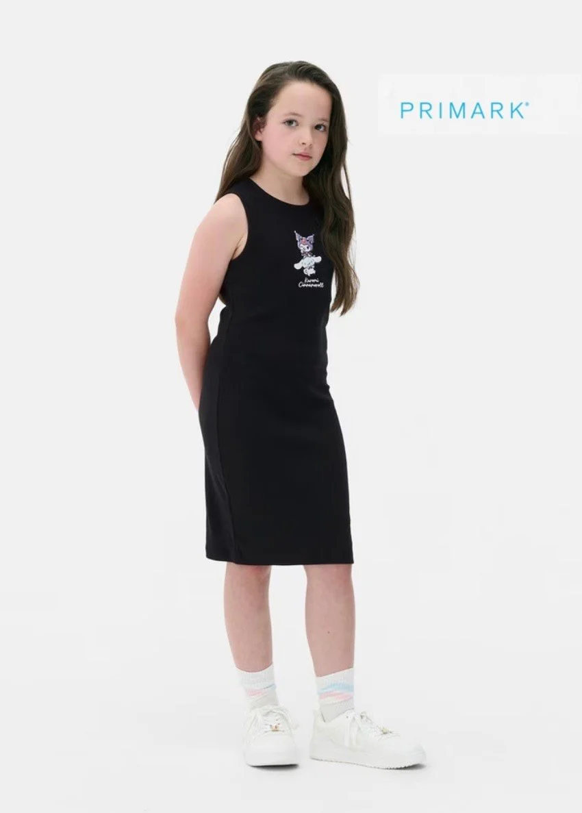 A young girl in a black sleeveless dress with a colorful animal graphic, white sneakers, and pastel striped socks, standing with her hands behind her back against a plain background, with the word 'PRIMARK' in blue text on the upper right.