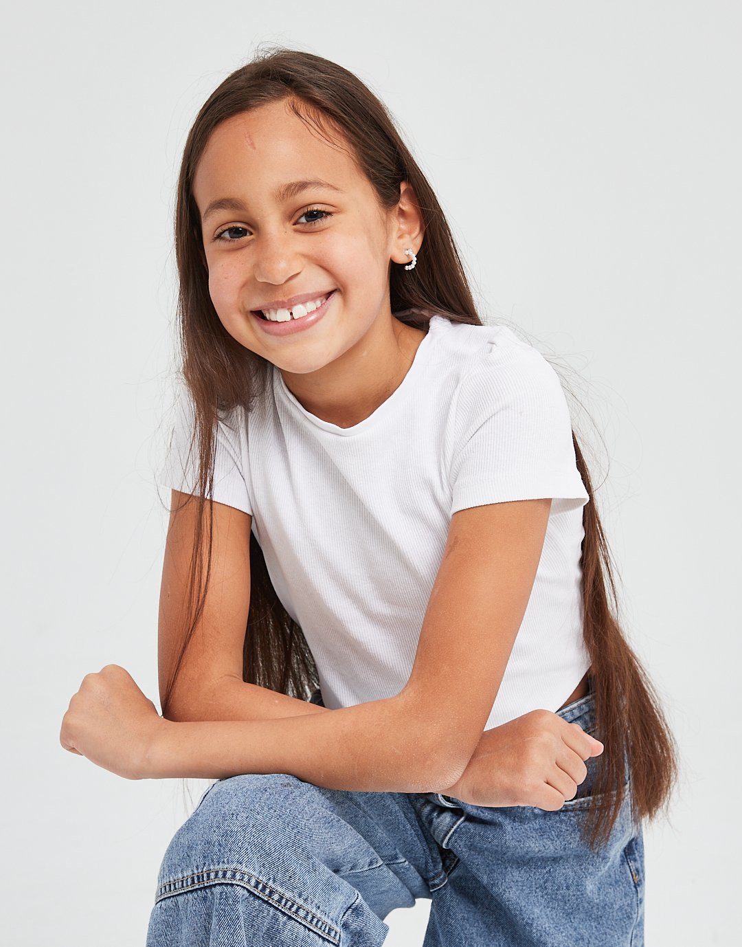 Smiling girl with long brown hair, wearing a white t-shirt and jeans, sitting against a plain white background.Modelling for leading child model and talent agency SocityUK based in London and Manchester. 