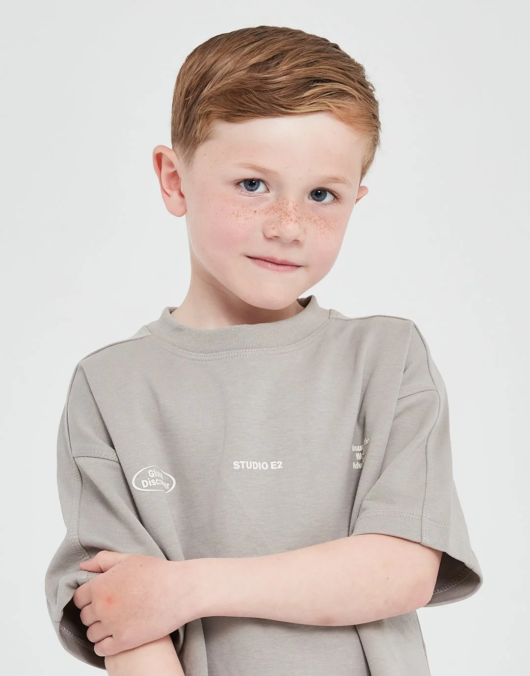 Young boy with red hair, blue eyes, and freckles, wearing a gray shirt with arms crossed, smiling softly at the camera against a plain white background. Modelling for leading child model and talent agency SocityUK based in London and Manchester.