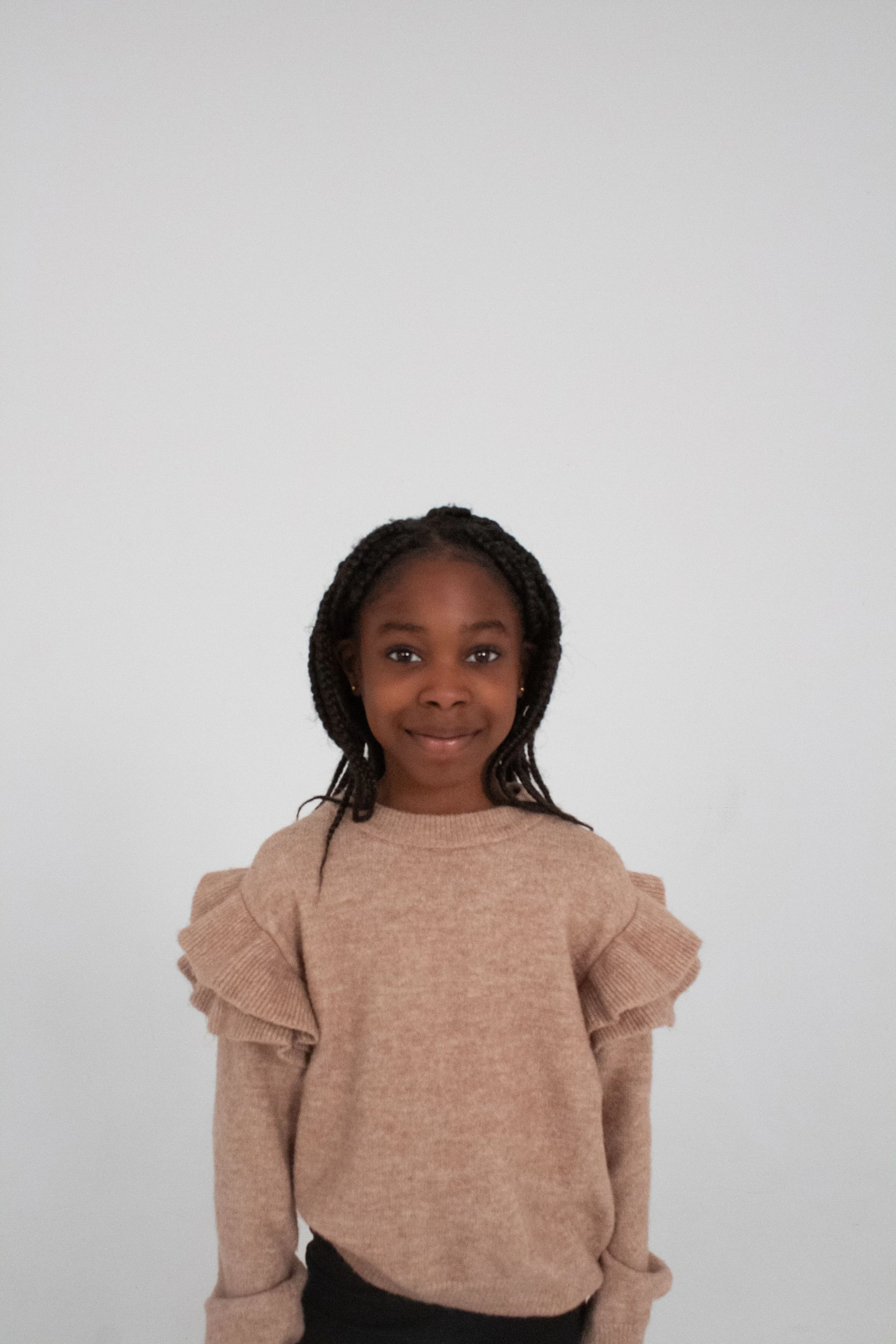 A young girl with braided hair smiling at the camera, wearing a beige sweater with ruffled shoulders, standing against a plain white background.