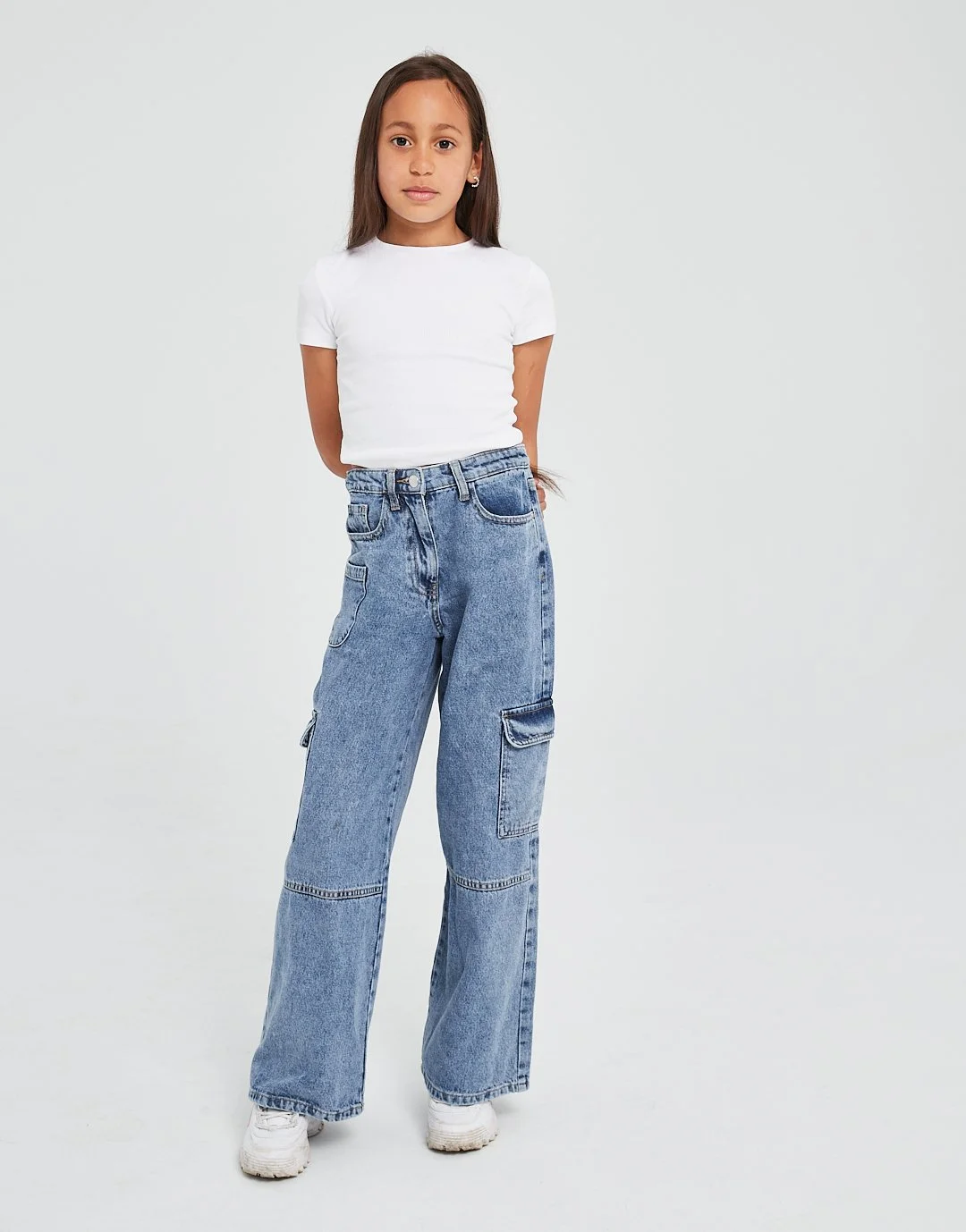 A young girl with long brown hair standing against a plain light background, wearing a white t-shirt, baggy blue jeans, and white sneakers. Modelling for leading child model and talent agency SocityUK based in London and Manchester.