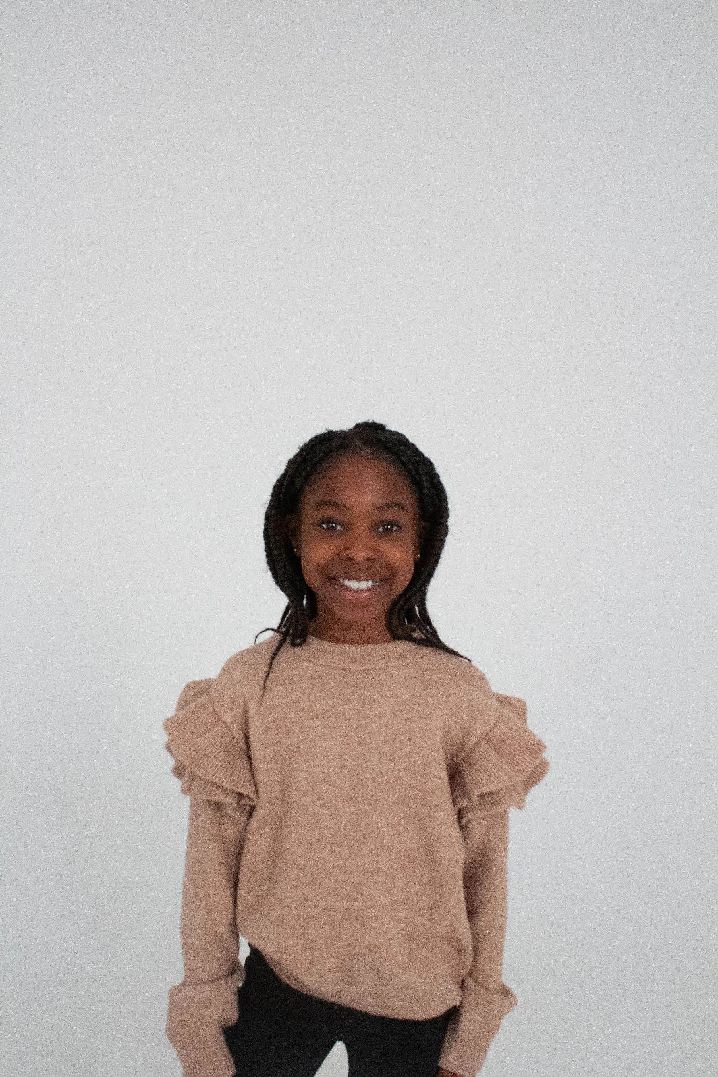 A young girl with braided hair smiling and standing in front of a plain white wall, wearing a beige sweater with ruffled shoulders.