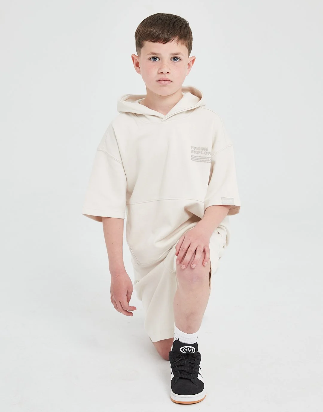 Young boy with brown hair and blue eyes in a loose, cream-colored hoodie and matching shorts, kneeling on one knee with one hand on his knee and the other by his side, against a plain white background.