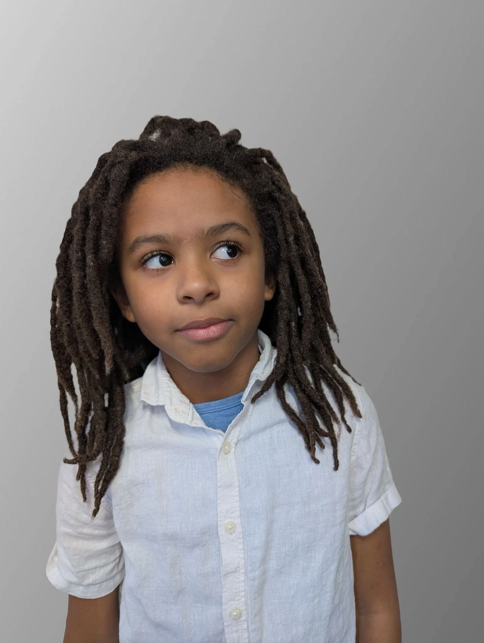 Young boy with long dreadlocks wearing a white shirt and light blue T-shirt underneath, looking to the side against a plain grey background.