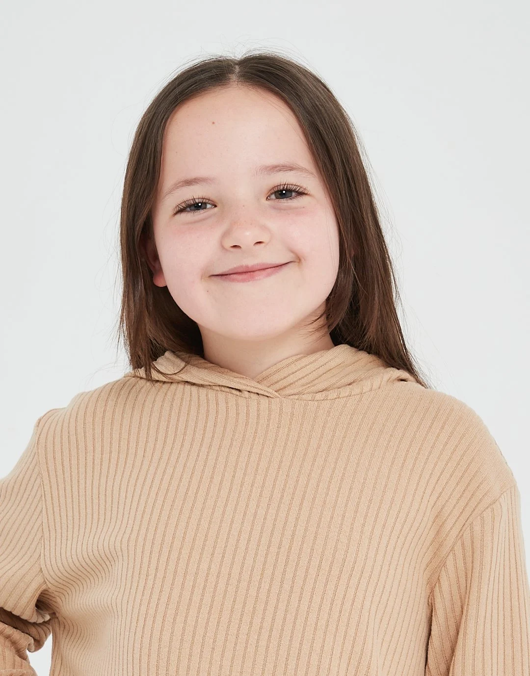 A young girl with long brown hair and a beige hoodie smiling at the camera against a plain white background.