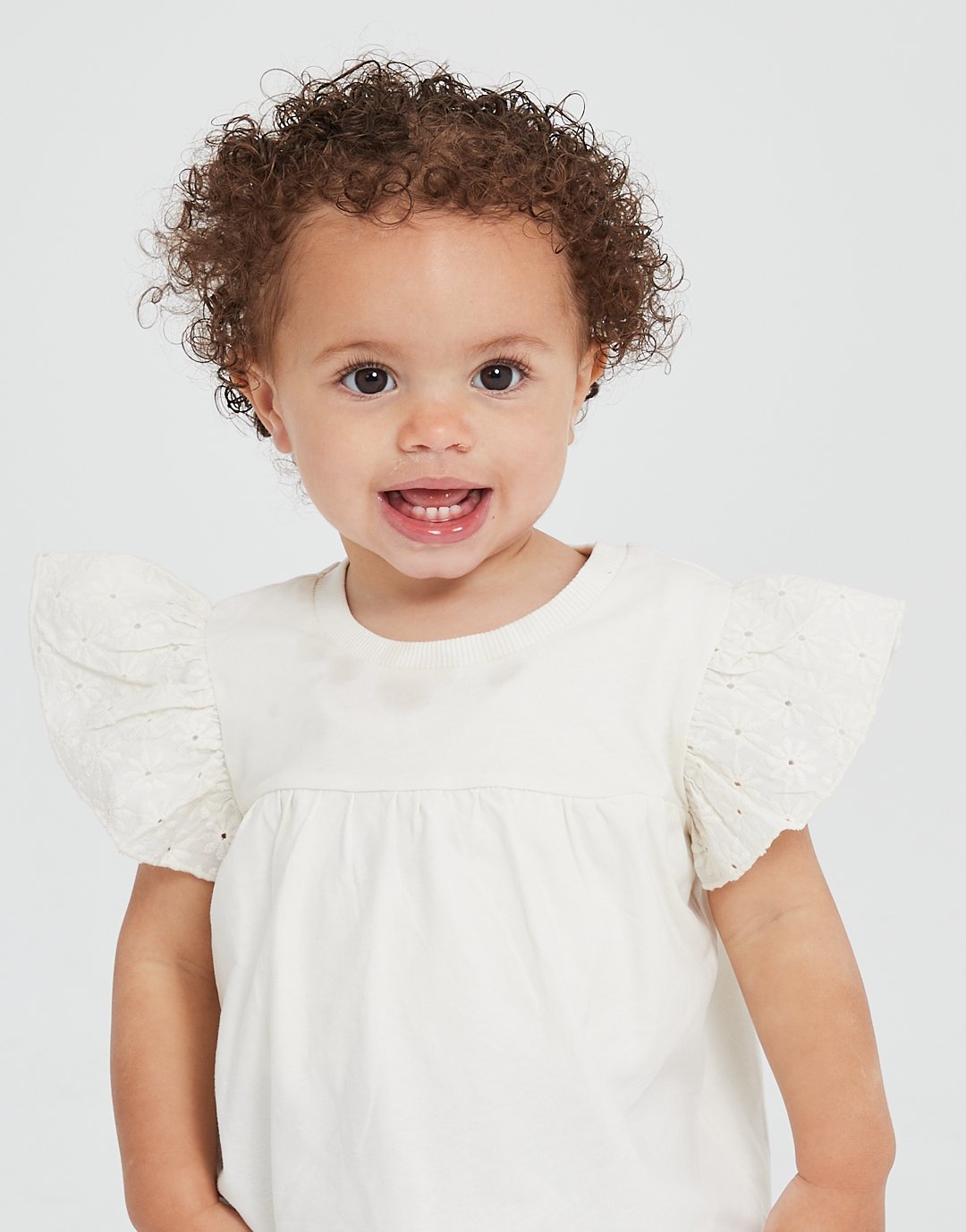 A young girl with curly hair and big blue eyes smiling, wearing a white dress with puffed sleeves. Modelling for leading child model and talent agency SocityUK based in London and Manchester.