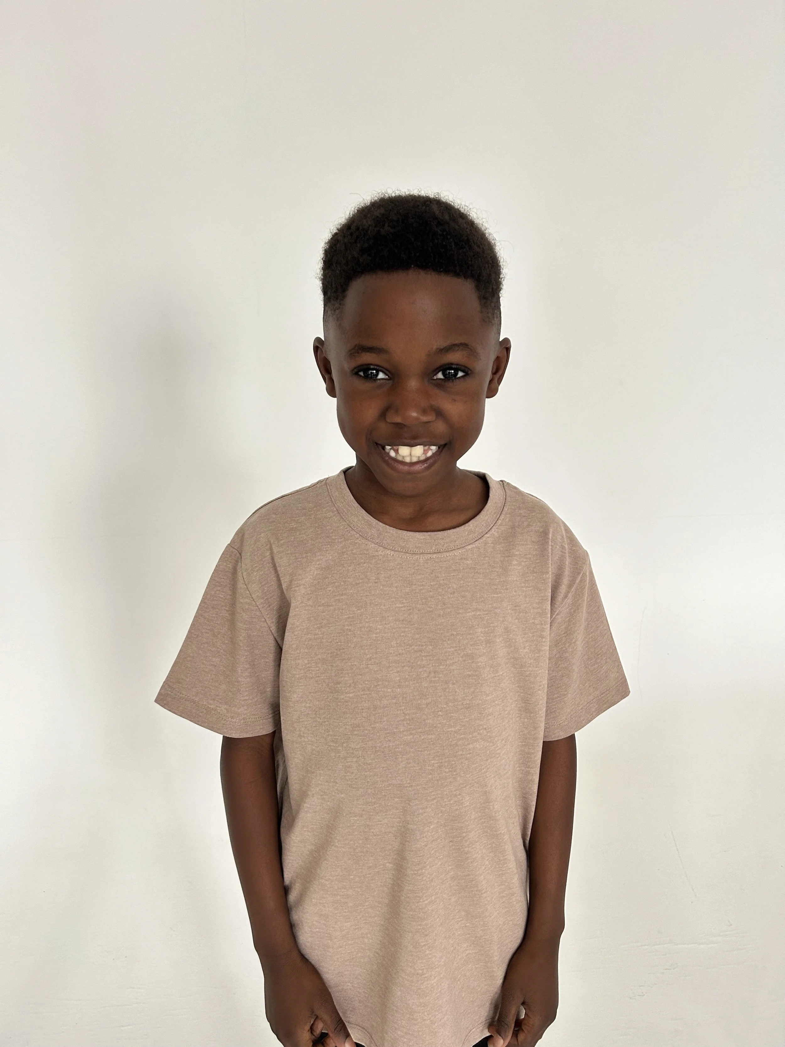 A young boy with a short afro hairstyle smiling, wearing a beige t-shirt, standing in front of a plain white background. Model for leading Manchester and London based child model agency Socityuk.