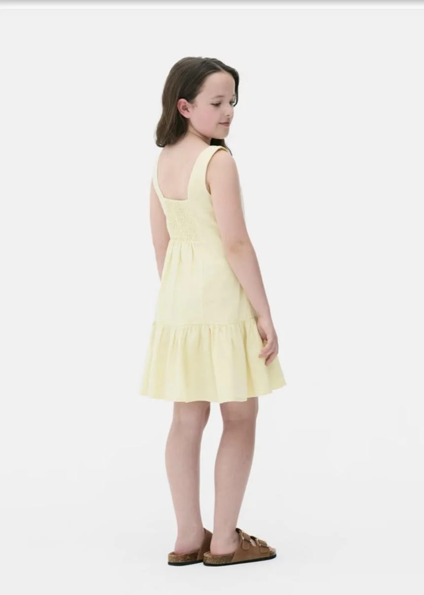 A young girl in a yellow sleeveless dress with brown sandals standing against a plain white background.