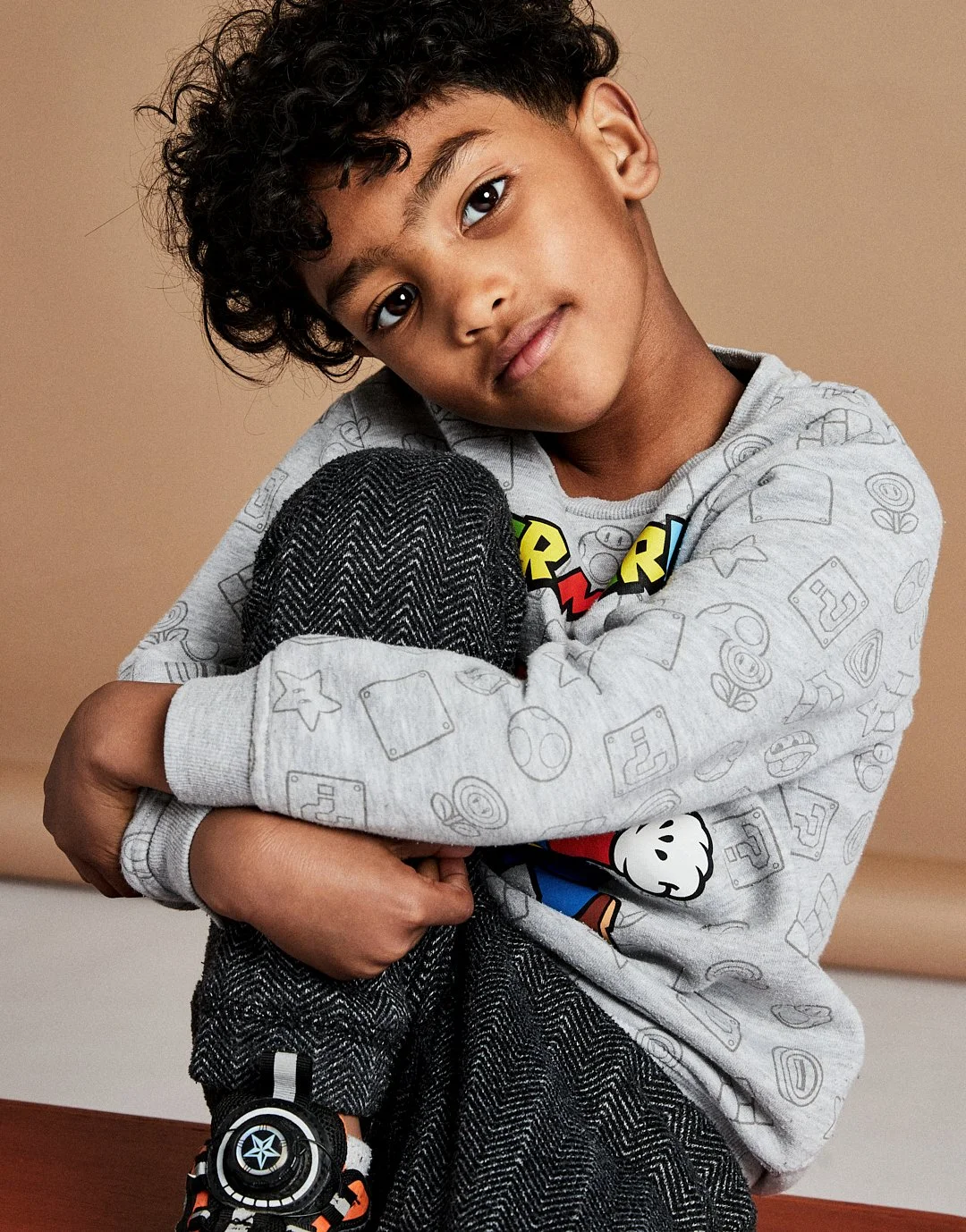A young boy with curly hair hugging his knee, wearing a light gray Mario-themed sweatshirt and dark gray herringbone pants, against a neutral background.