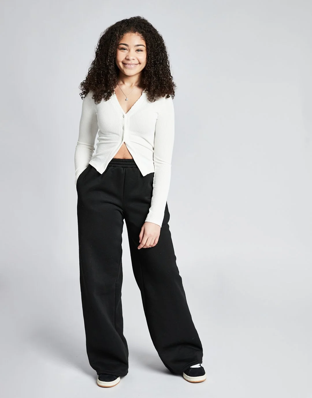 A young woman with curly hair standing against a light gray background, smiling, wearing a white cropped button-up sweater, black loose-fitting pants, and black sneakers.