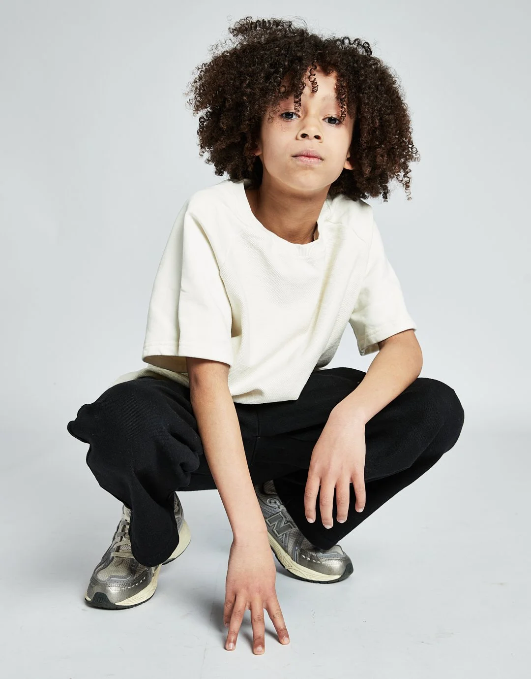 A young boy with curly hair in a squatting pose, wearing a white t-shirt, black pants, and athletic shoes, against a plain light gray background.