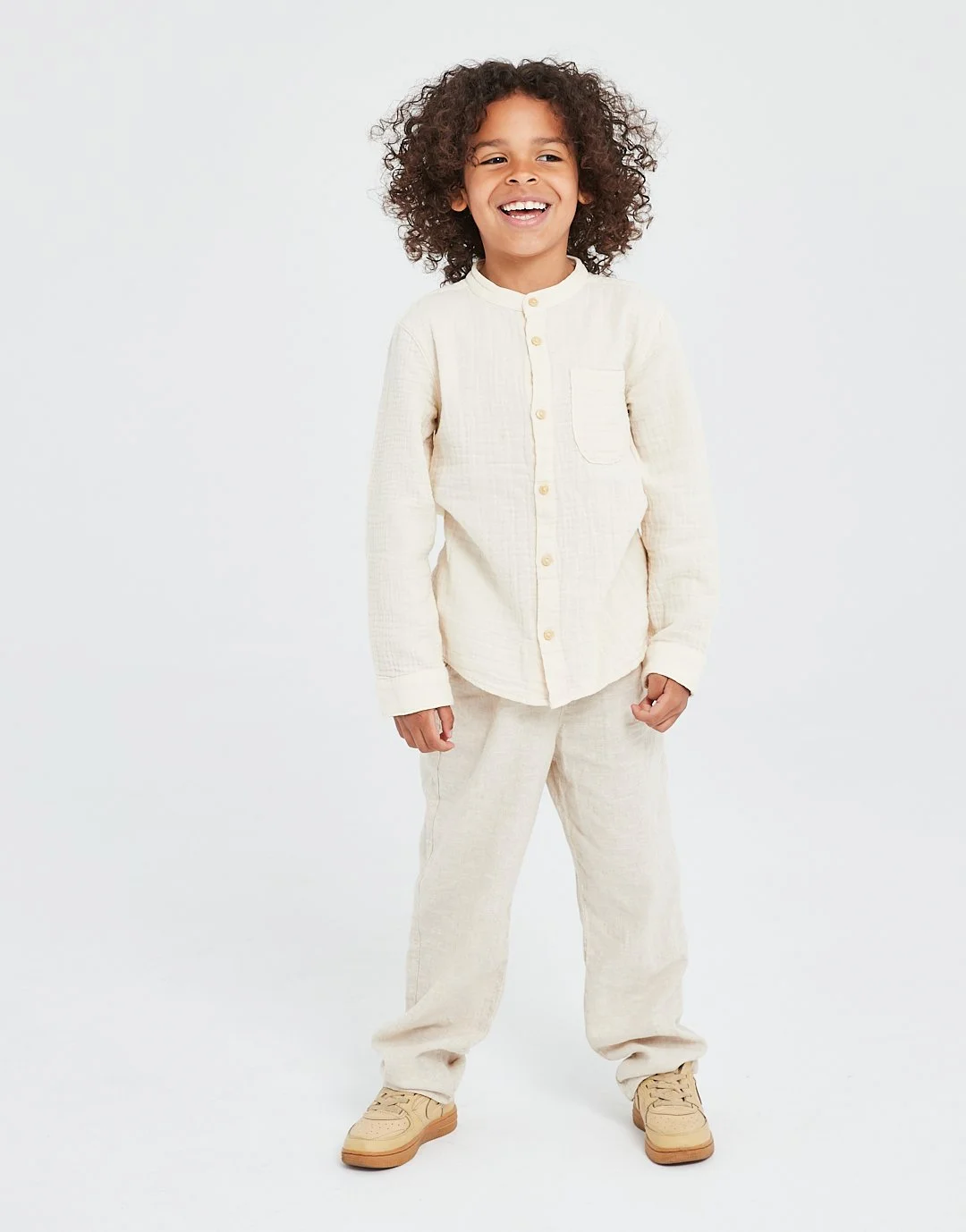 A young boy with curly hair smiling, dressed in light-colored pajamas and beige shoes, standing against a plain white background.