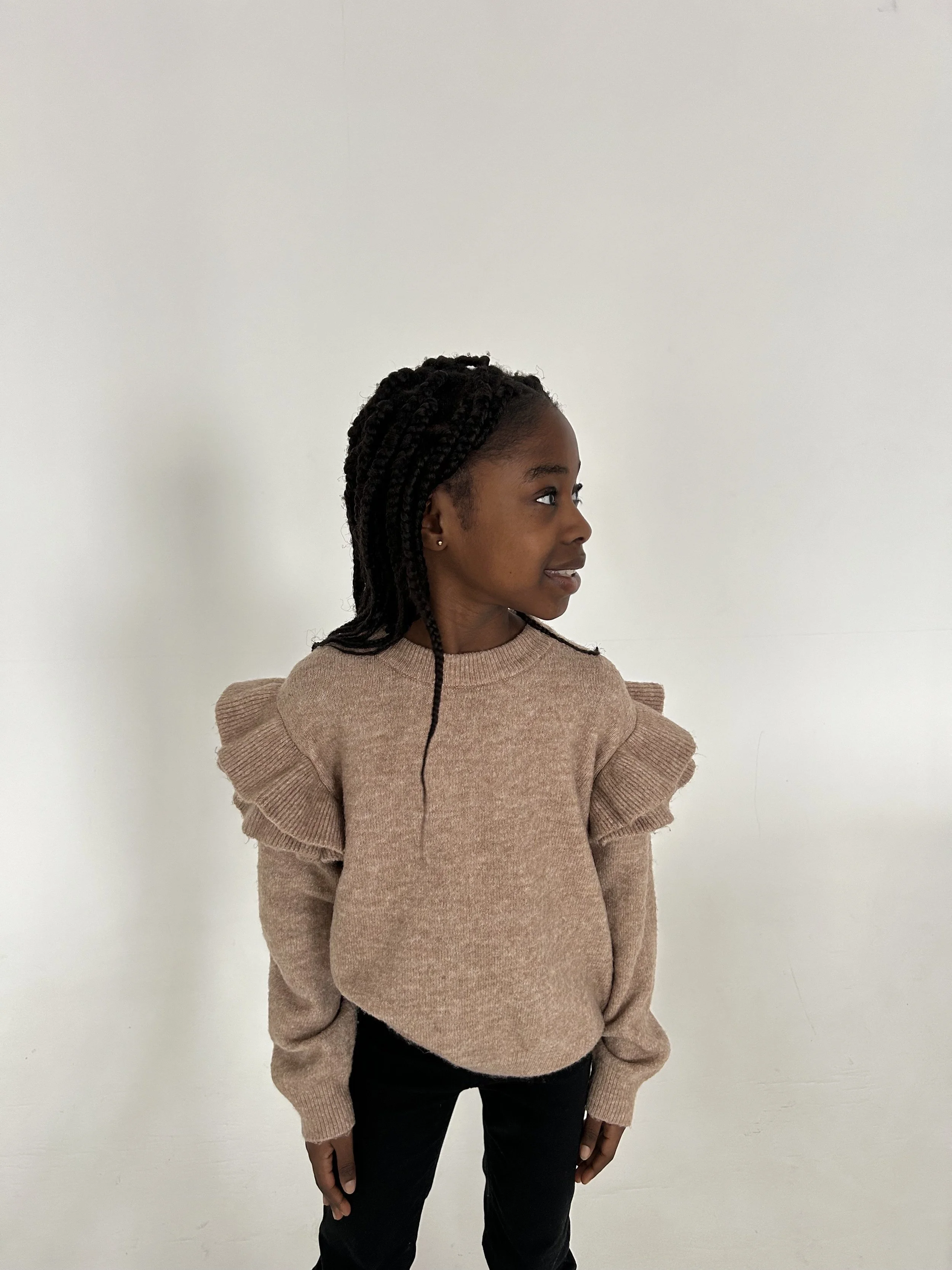 A young girl with dark braided hair wearing a beige sweater with ruffled shoulders and black pants, standing against a plain white wall, looking to her right.