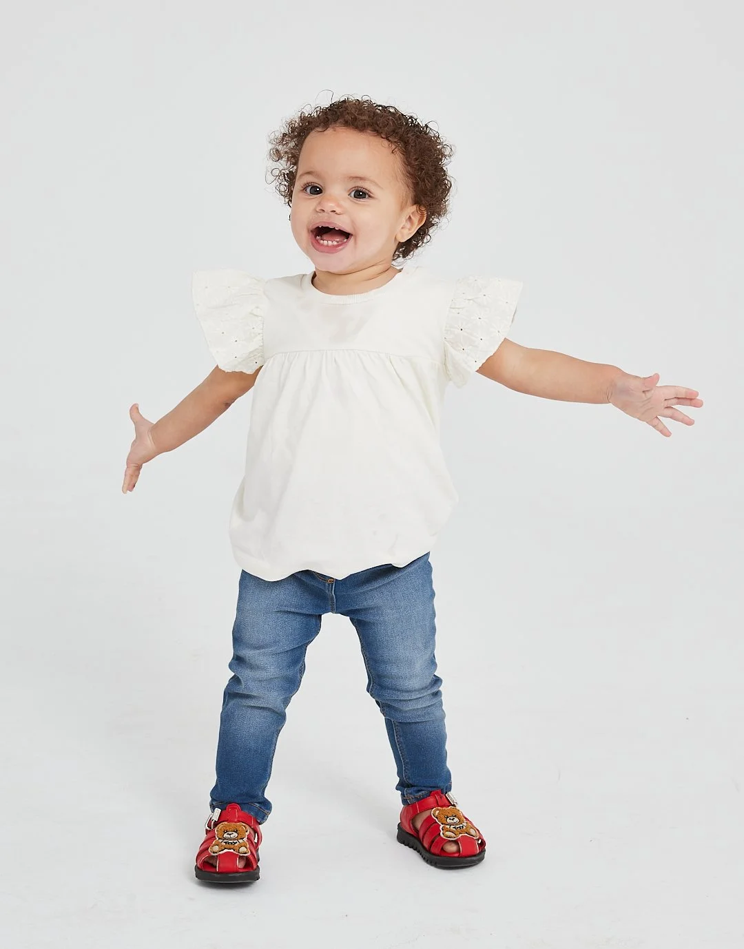 Young girl with curly hair smiling, arms outstretched, wearing a white top, blue jeans, and red sandals with teddy bear designs, standing against a plain white background. Modelling for leading child model and talent agency SocityUK based in London a