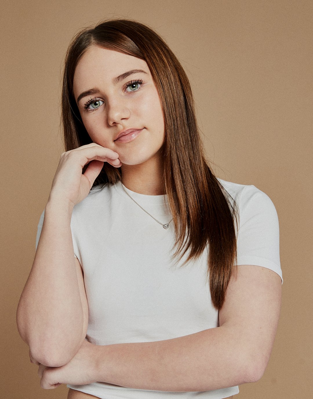 A young woman with long brown hair, blue eyes, and fair skin, wearing a white T-shirt and a necklace, touching her chin with her right hand and smiling softly in front of a beige background.