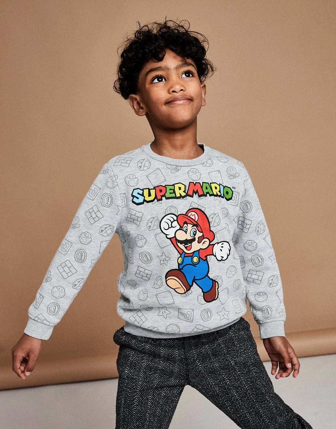 A young boy with curly dark hair wearing a light gray sweatshirt featuring a large picture of Super Mario, with the word 'Super Mario' in colorful letters above the character, and dark gray textured pants, standing against a tan background.