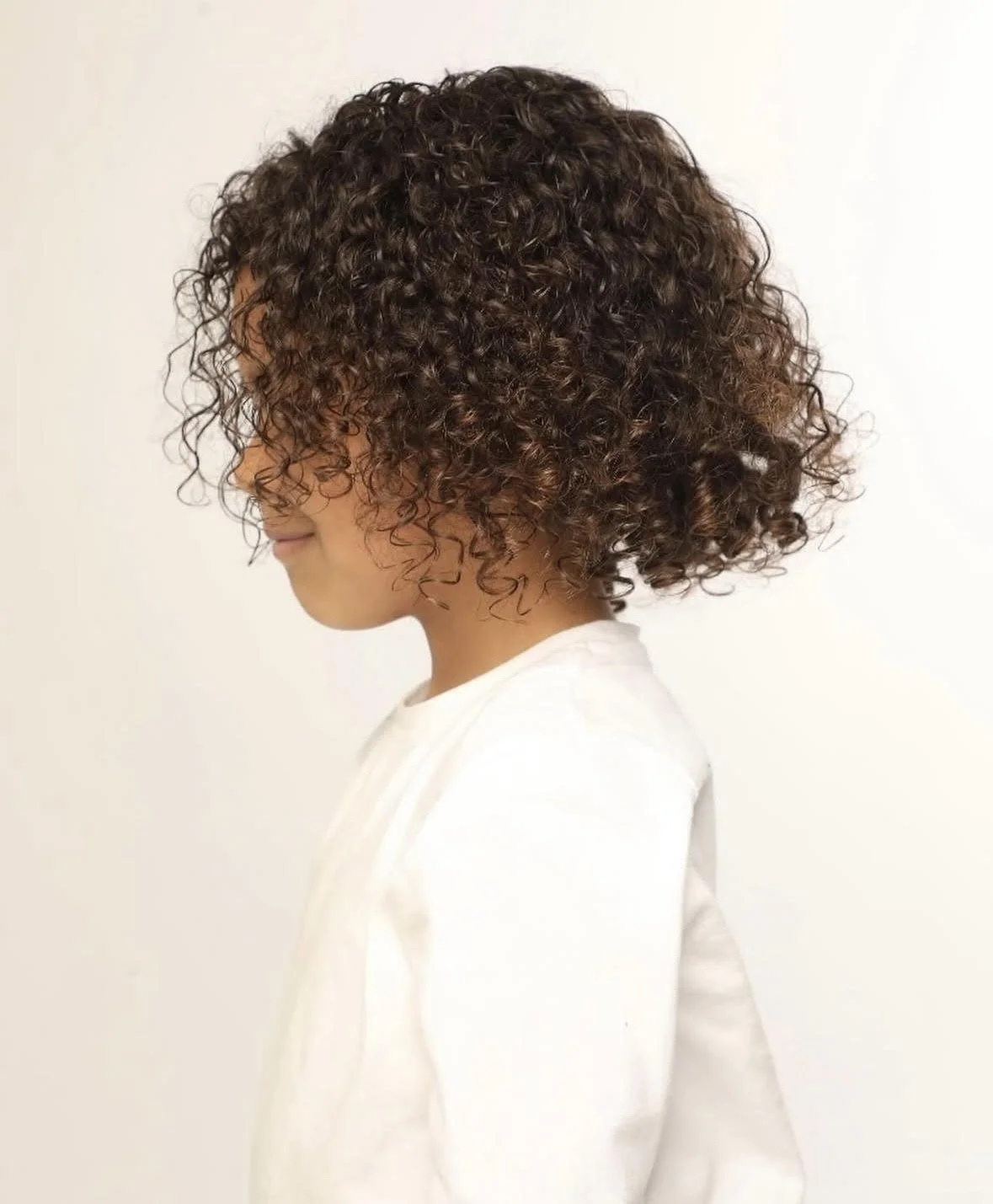 Side view of a child with short, curly brown hair, wearing a white hoodie, standing against a plain white background.
