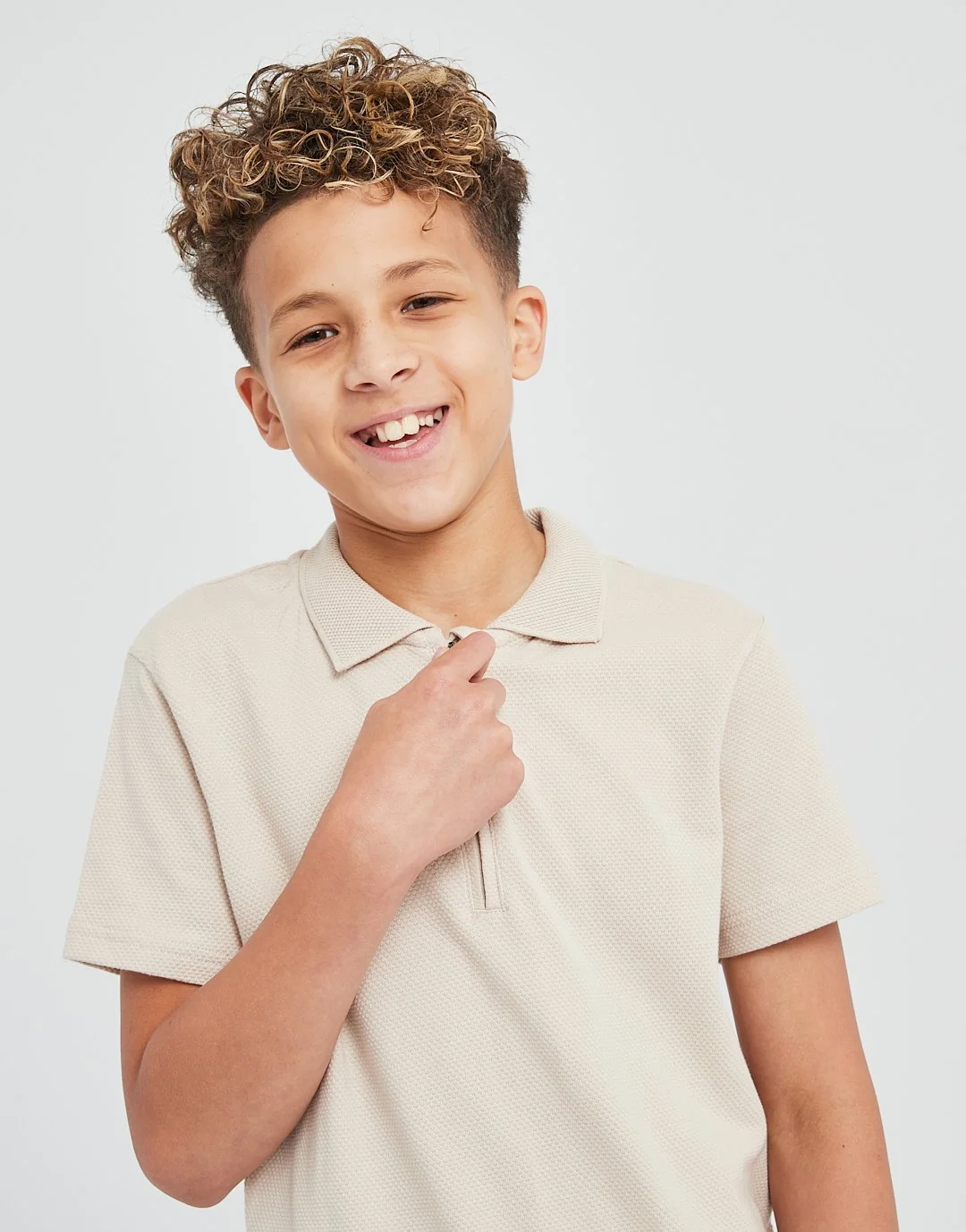 A boy with curly light brown hair smiling and holding his hand near his neck, wearing a beige polo shirt, against a plain light background.