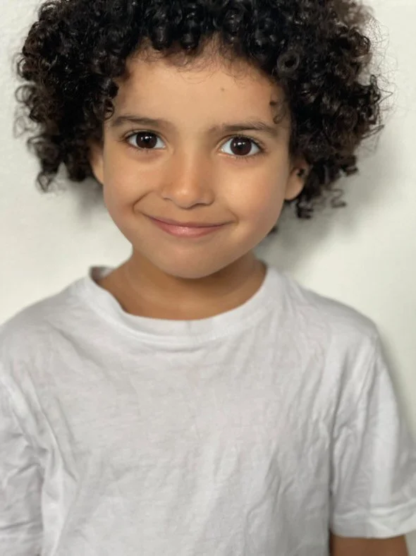 A young girl with curly hair, smiling, wearing a white shirt against a plain background.