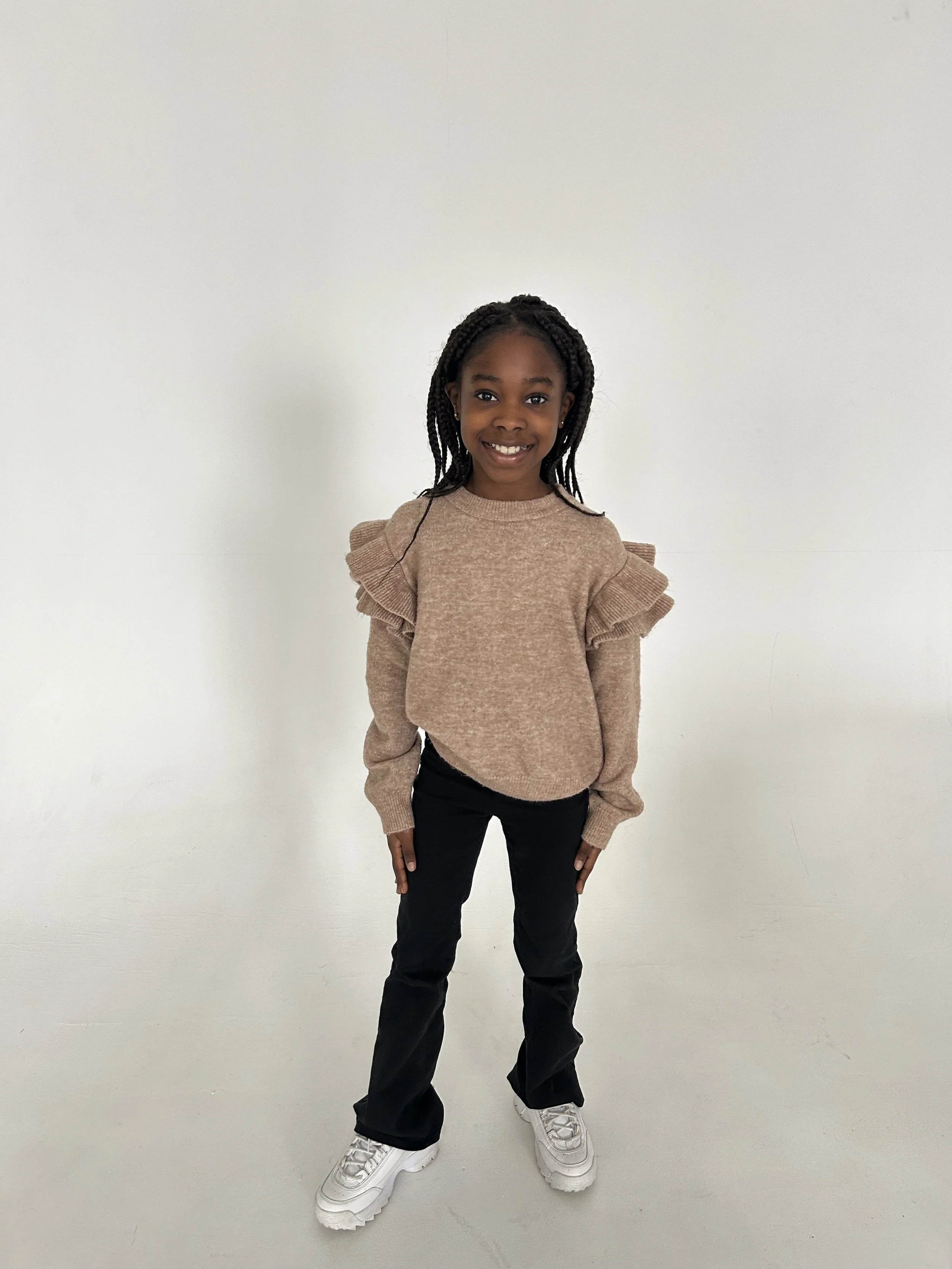 A young girl with braided hair standing and smiling against a plain white wall, wearing a beige sweater with ruffled shoulders, black pants, and white sneakers. Model for leading Manchester and London based child model agency Socityuk.