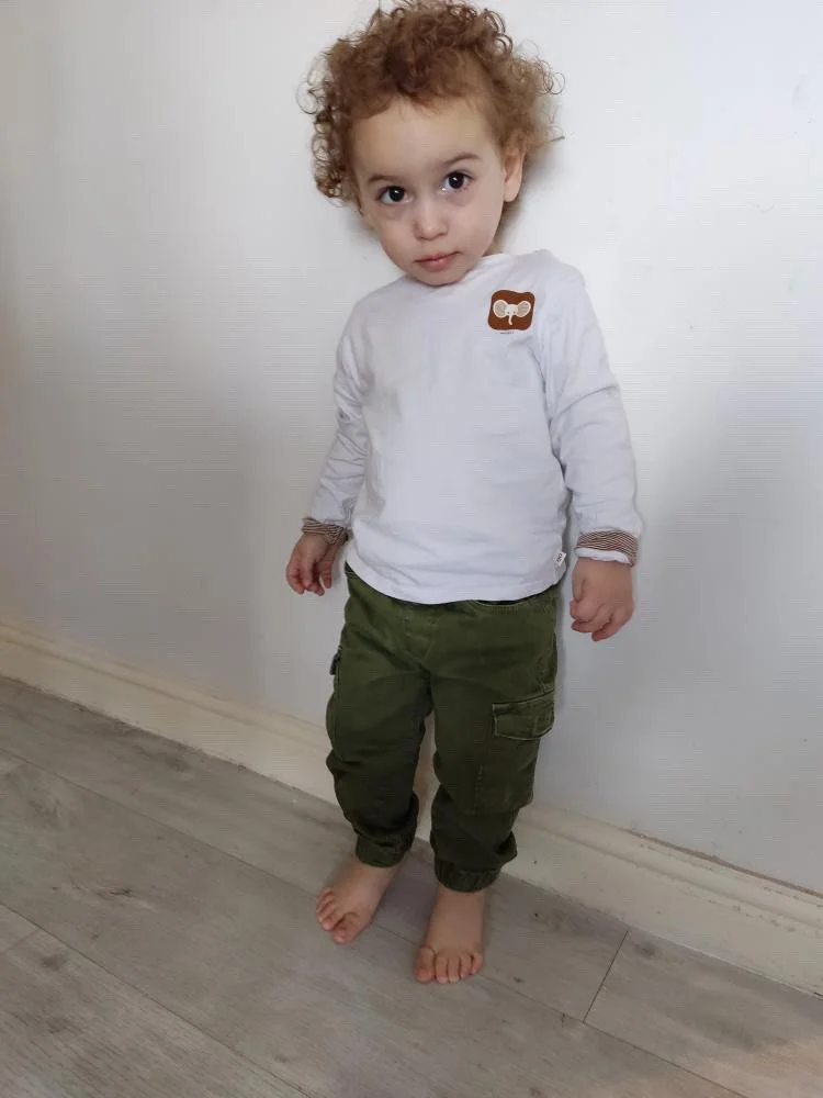A young boy with curly hair standing barefoot against a plain white wall, wearing green cargo pants and a long-sleeve white shirt with a small brown bear face patch.