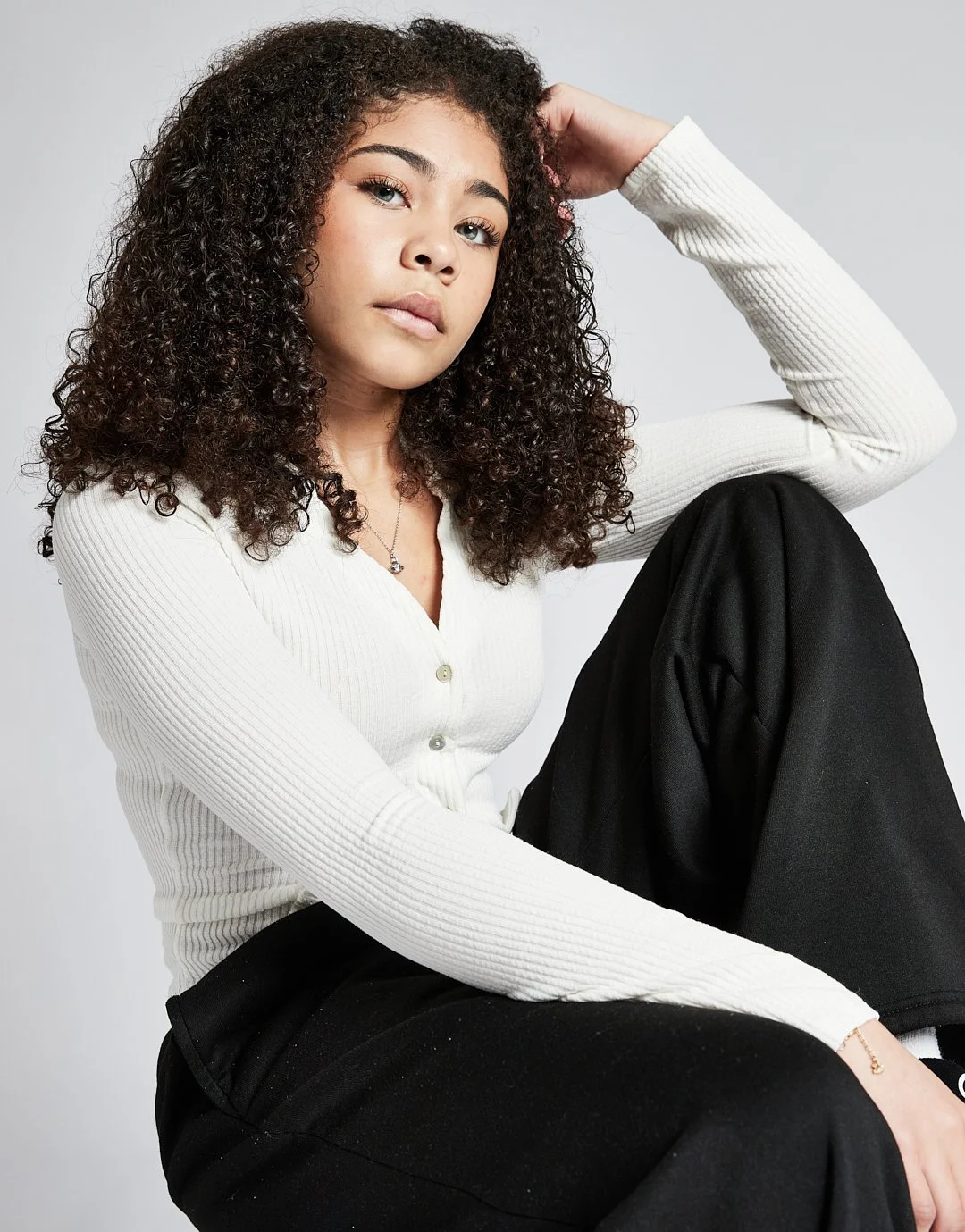 A young woman with curly hair wearing a white long-sleeve shirt, sitting with one knee up, looking at the camera against a plain light gray background.