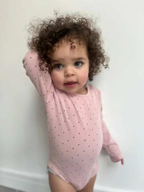 A young girl with curly hair and blue eyes wearing a pink polka dot long-sleeve outfit standing against a plain white wall. Modelling for leading child model and talent agency SocityUK based in London and Manchester.