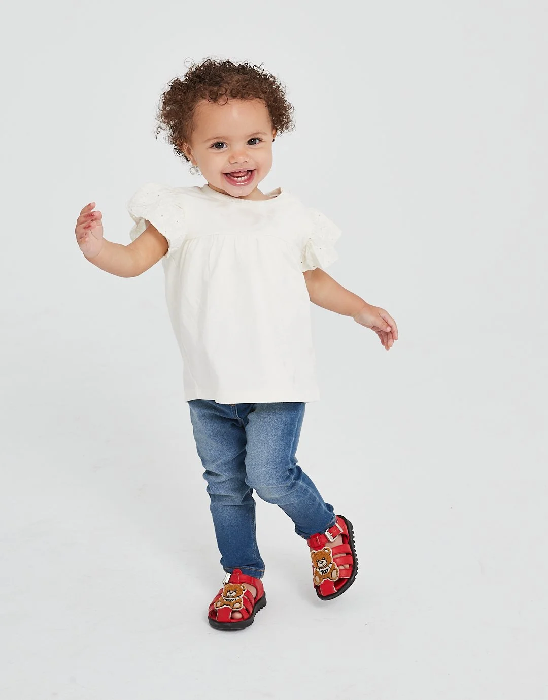 A young girl with curly hair smiling and dancing, wearing a white top, blue jeans, and red sandals with teddy bear decoration, standing against a plain white background. Modelling for leading child model and talent agency SocityUK based in London and