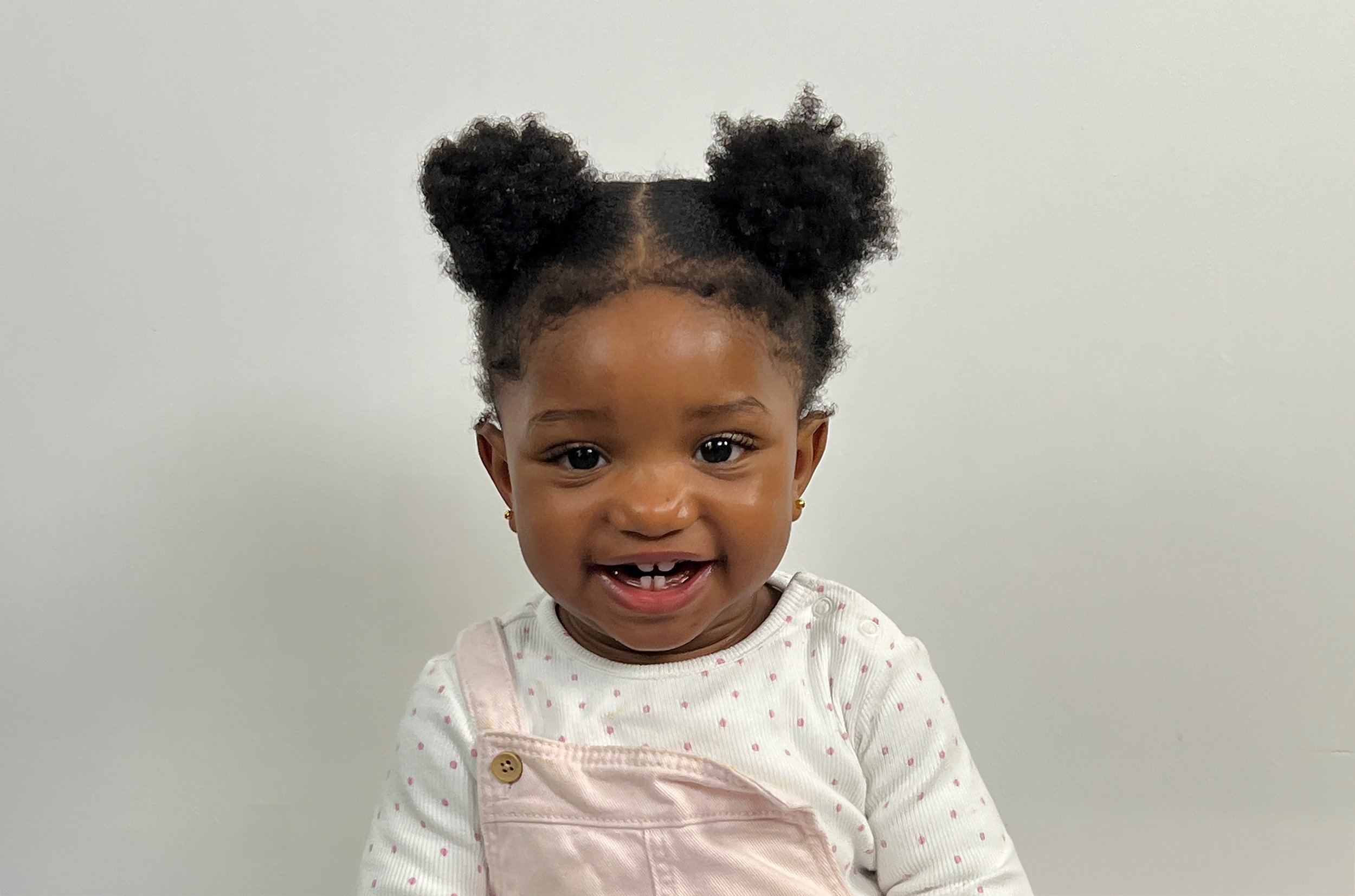 A young girl with curly hair styled in puffs, smiling and showing her teeth, wearing a white shirt with pink polka dots and light pink overalls, standing against a plain white wall.
